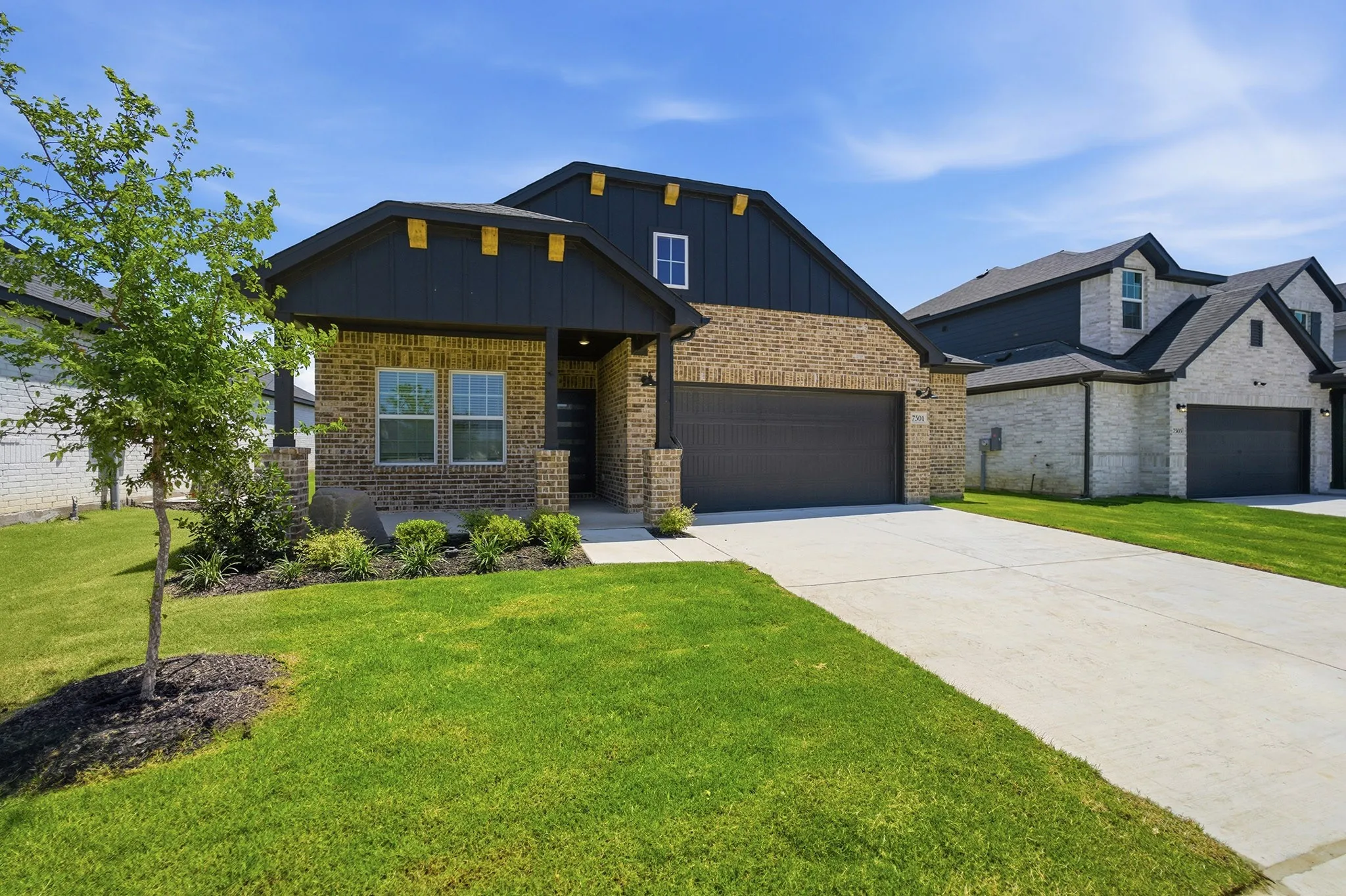 Contemporary residence featuring a brick and dark siding exterior, an attached garage, a covered front porch, a concrete driveway, and a landscaped front yard with a young tree