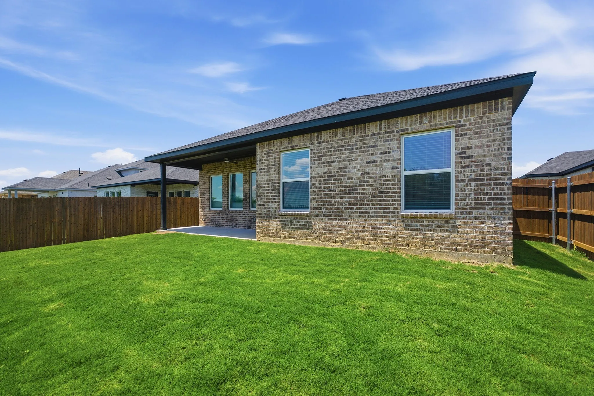 Expansive green lawn with a covered patio, brick exterior, dark shingle roof, and privacy fencing