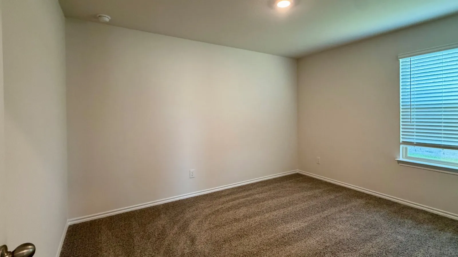 Recessed ceiling lighting illuminates a room featuring a window with horizontal blinds, neutral wall paint, and brown carpet flooring