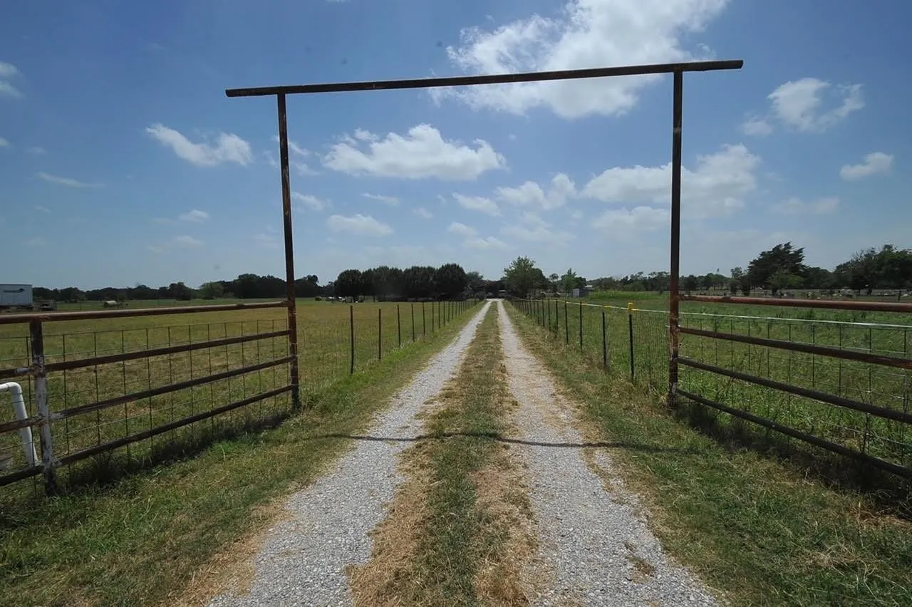 Gravel driveway with metal frame entrance, flanked by fencing and open fields