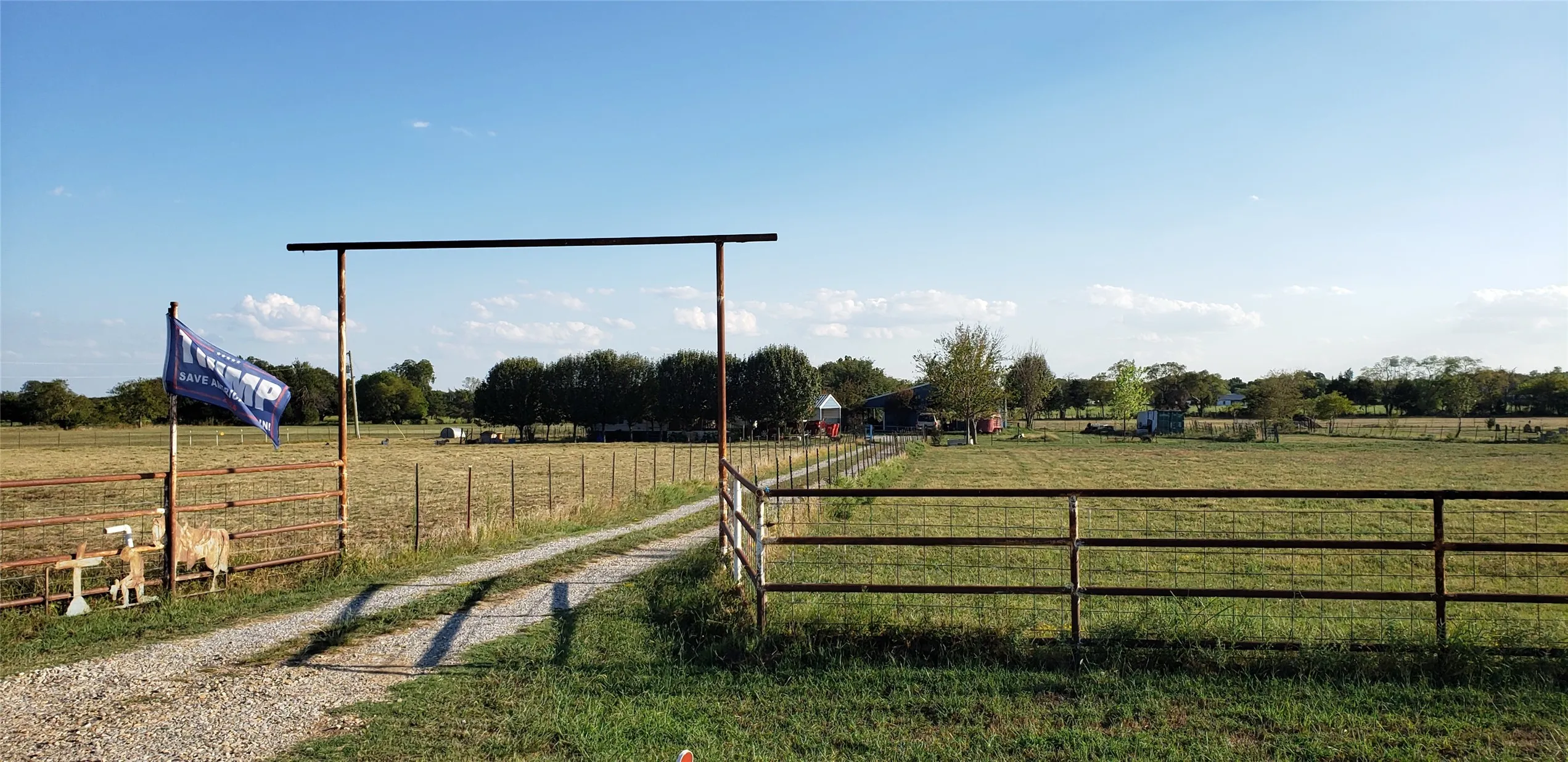 Gravel driveway entrance with a metal gate frame, leading to expansive fenced pastures and distant structures