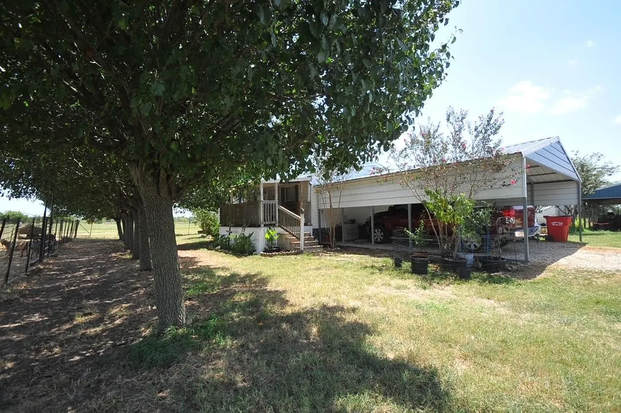 Exterior featuring an expansive metal carport, a raised wooden deck with railings, and a row of mature trees bordering a fenced perimeter