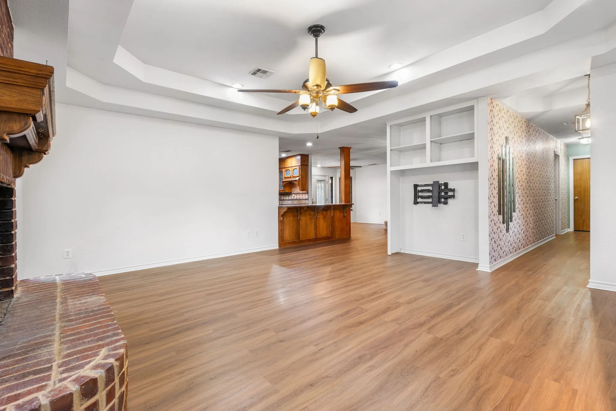 Spacious living area featuring wood-finish flooring, a brick fireplace with a wood mantel, and a tray ceiling with a ceiling fan