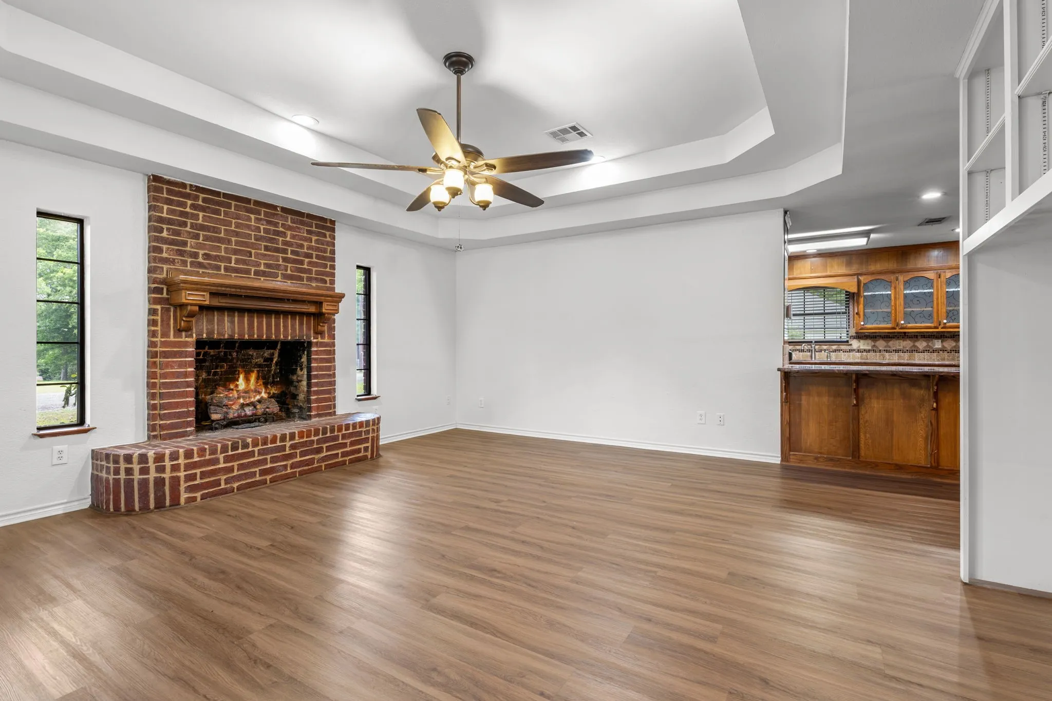 Spacious room featuring a brick fireplace with wood mantel, wood-finish flooring, and a tray ceiling with a fan