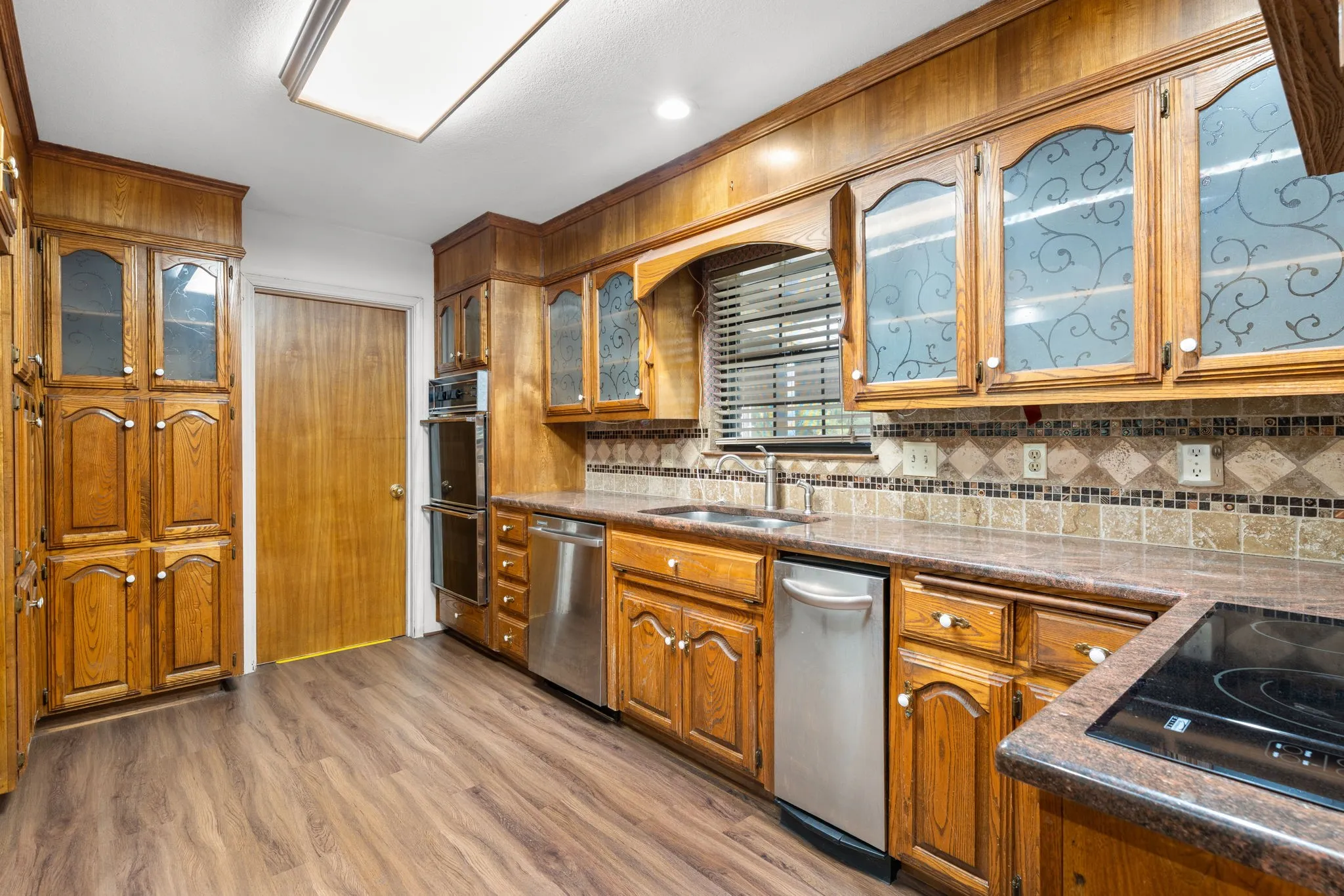 Kitchen featuring extensive wood cabinetry, stainless steel appliances, patterned glass cabinet doors, a mosaic tile backsplash, and wood-finish flooring