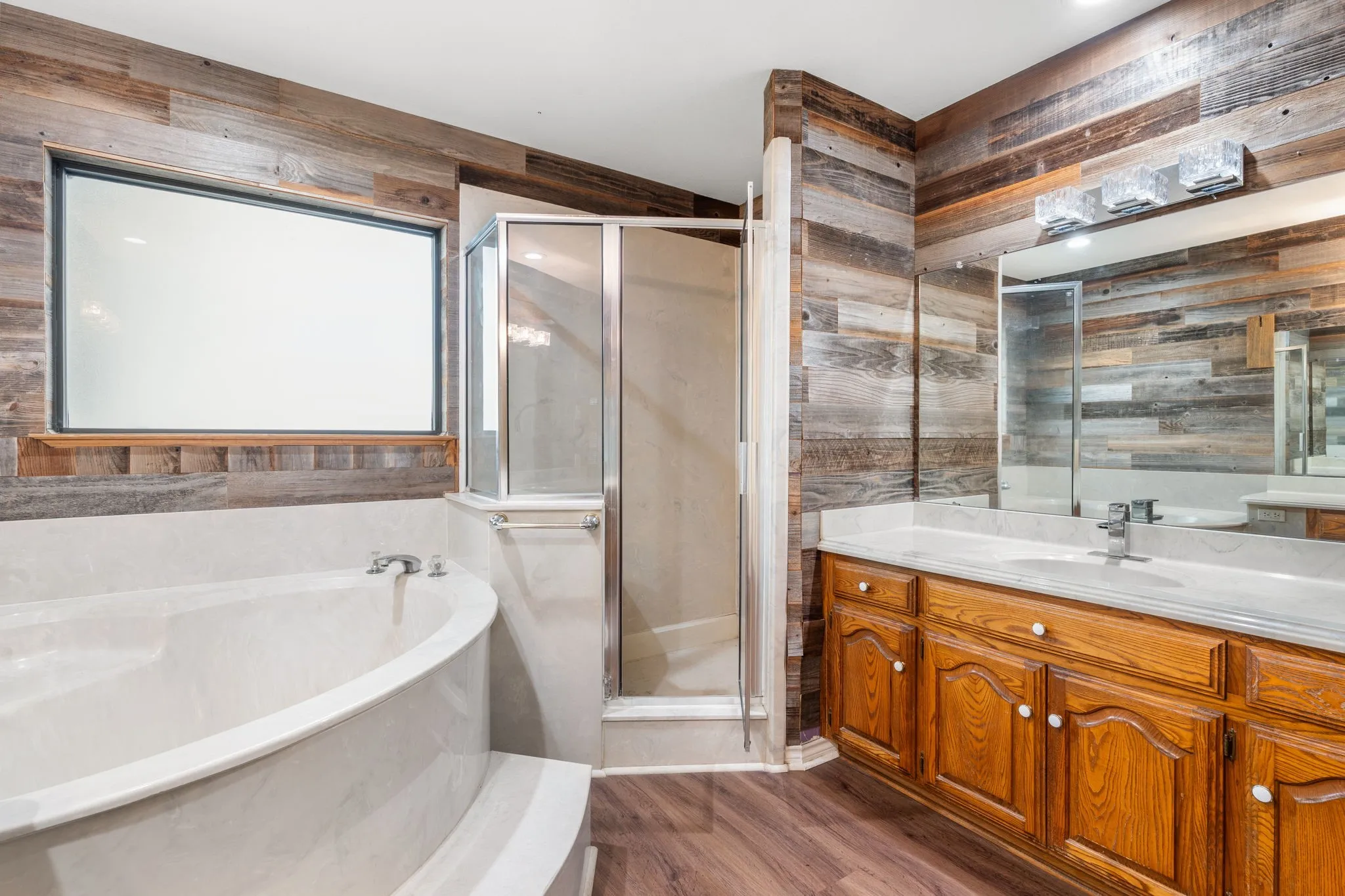 Bathroom featuring wood-finish flooring, a wood-paneled accent wall, a built-in soaking tub with a tiled surround, a glass-enclosed shower, and a dual-sink vanity with wood cabinetry