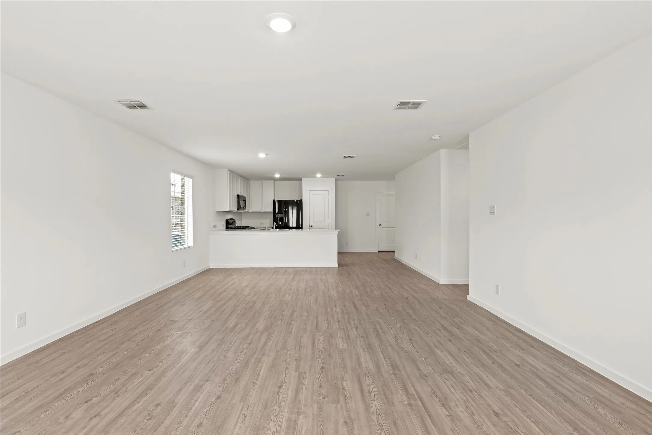 Spacious open-concept interior featuring light wood-finish flooring, recessed lighting, and a kitchen with white cabinetry, dark countertops, and black appliances