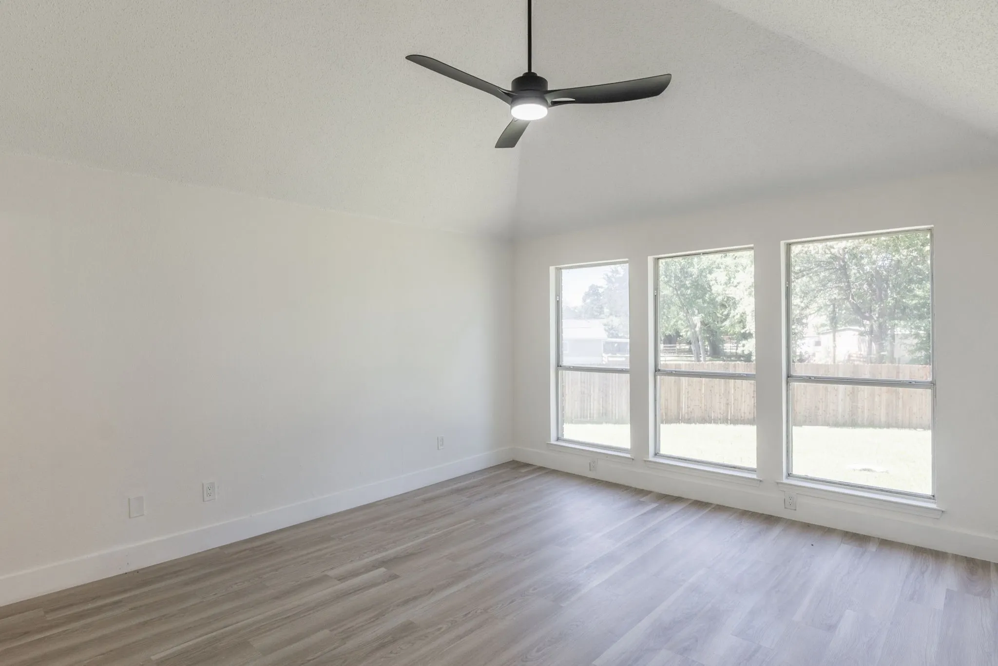 Room with a vaulted ceiling, a modern ceiling fan, three large windows, and light wood-finish flooring