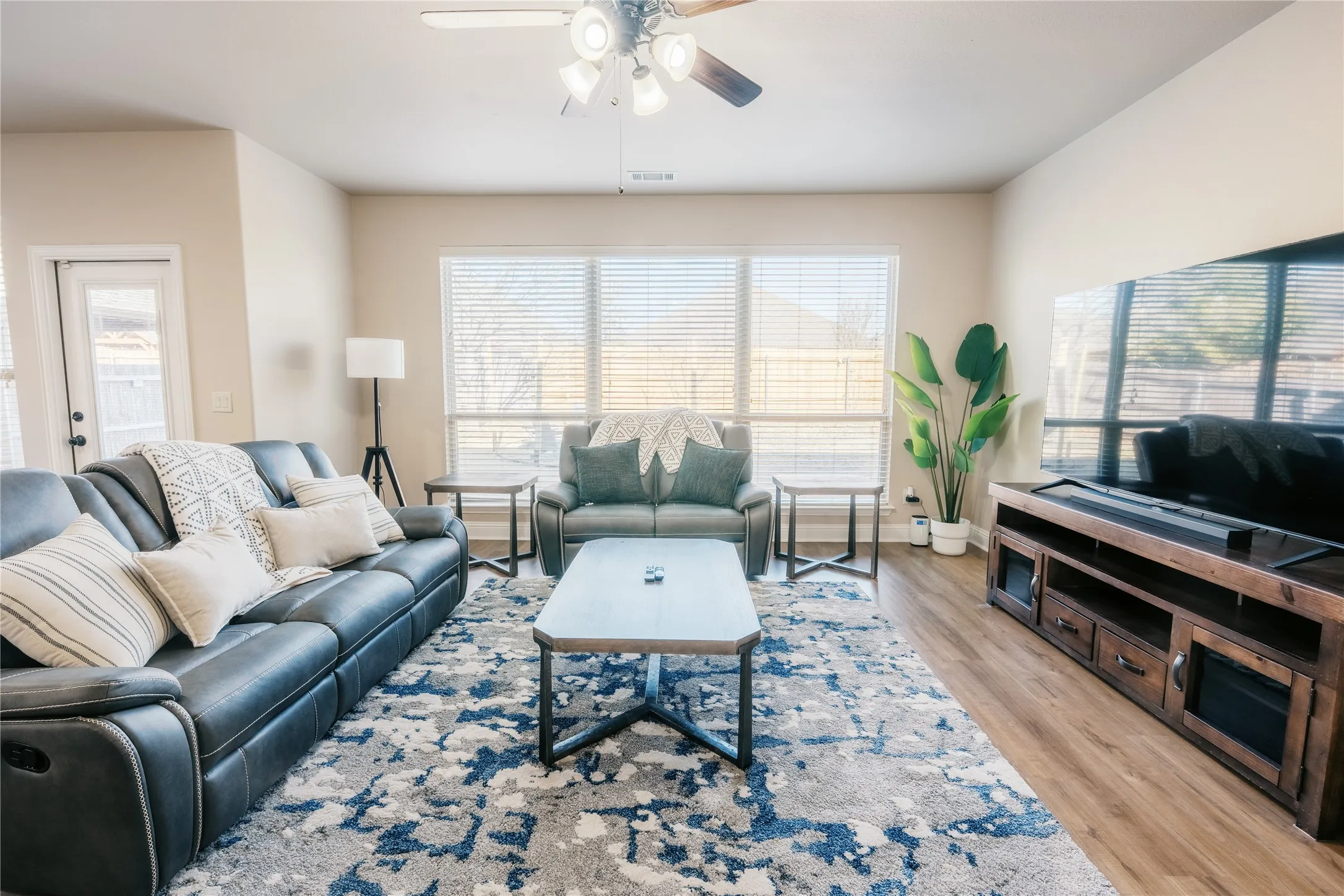 Spacious living area featuring wood-finish flooring, ample natural light from large windows, a built-in ceiling fan with lighting, and a rear door providing exterior access