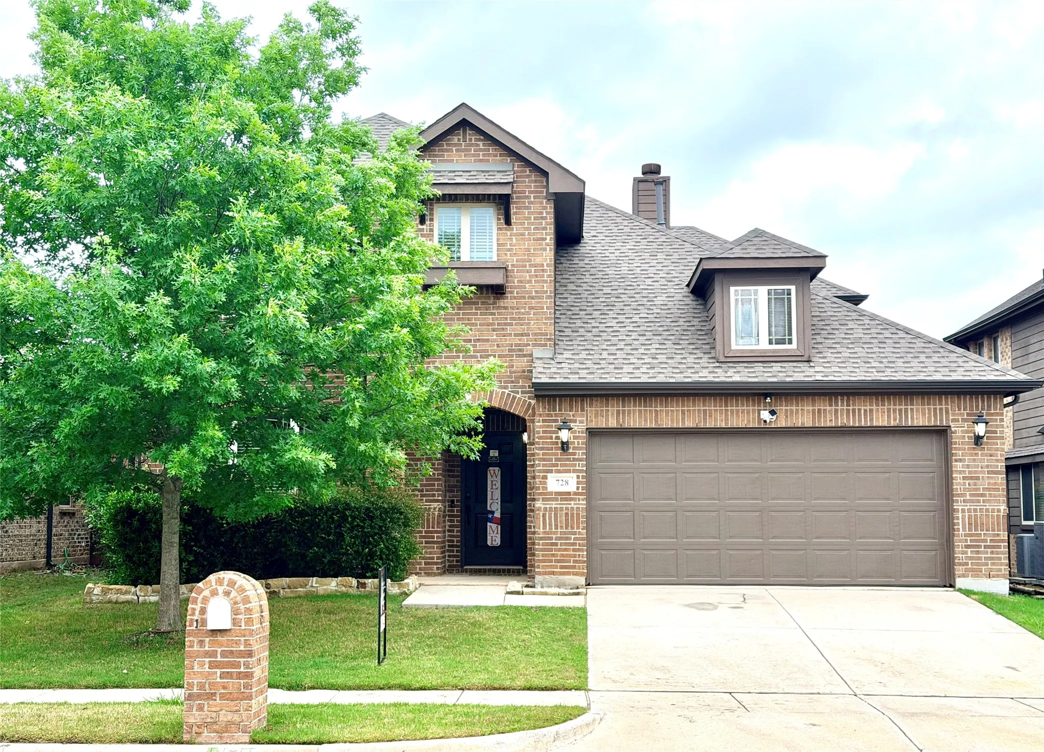 Brick facade residence featuring an attached two-car garage with a brown panel door, an arched entryway, dormer windows, and a shingle roof