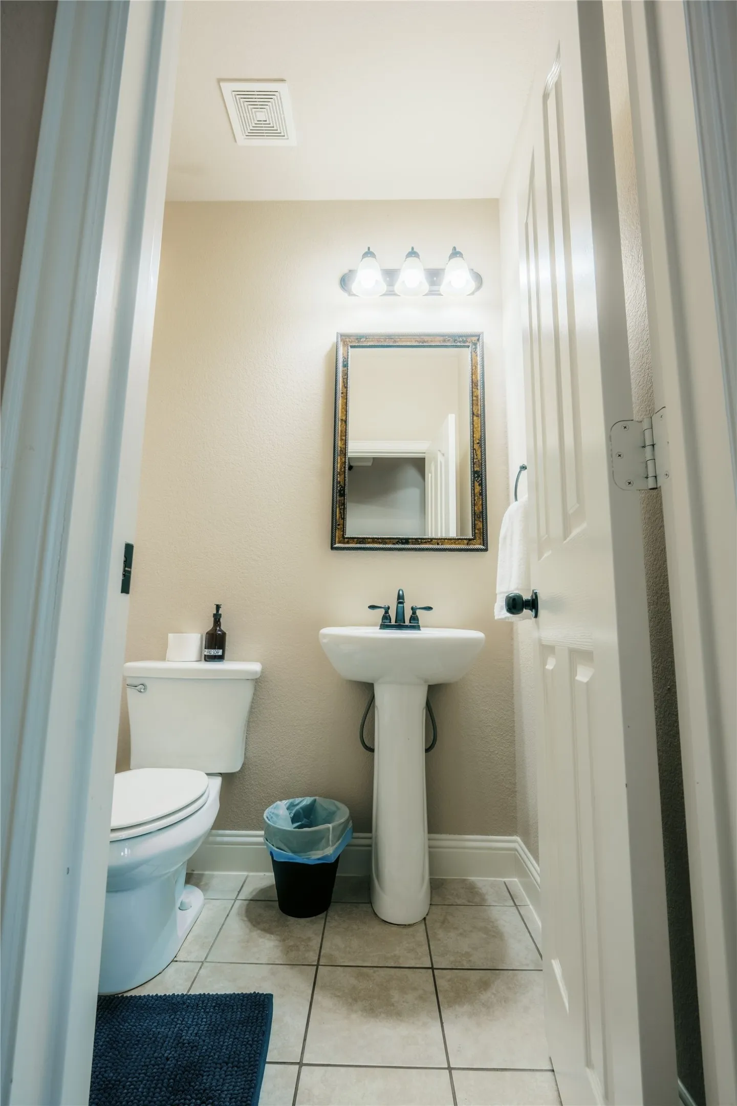 Compact bathroom featuring neutral-toned walls, tile flooring, a pedestal sink with a dark-finish faucet, a white toilet, and a decorative framed mirror with a three-bulb vanity light fixture