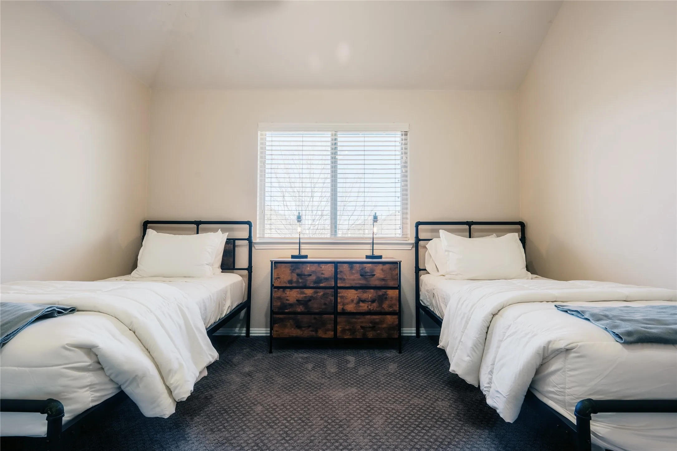 Vaulted ceiling room featuring a window with blinds, dark patterned carpeting, and painted neutral walls
