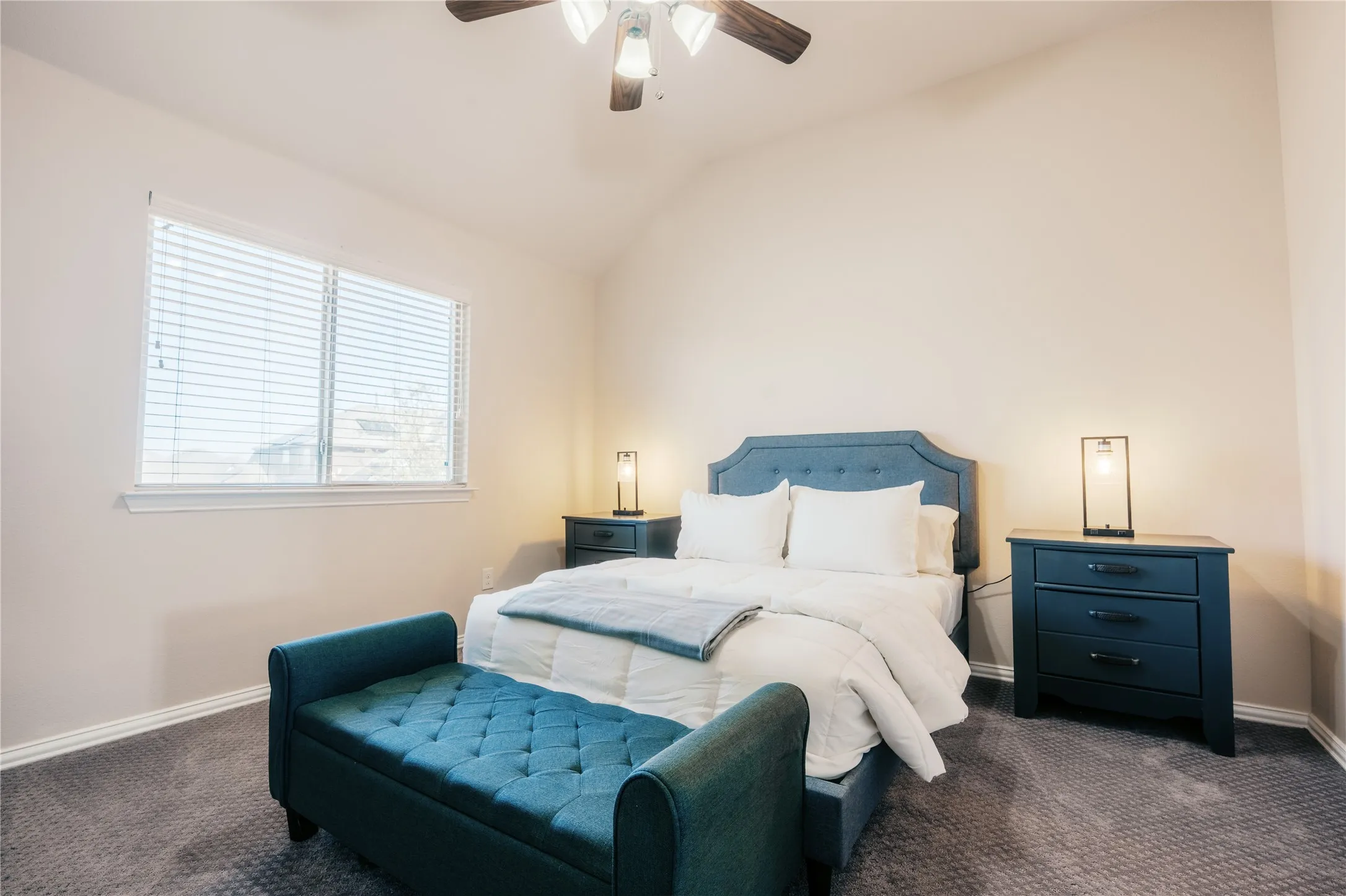 Carpeted bedroom featuring a vaulted ceiling with a ceiling fan, a large window with horizontal blinds, and neutral wall tones