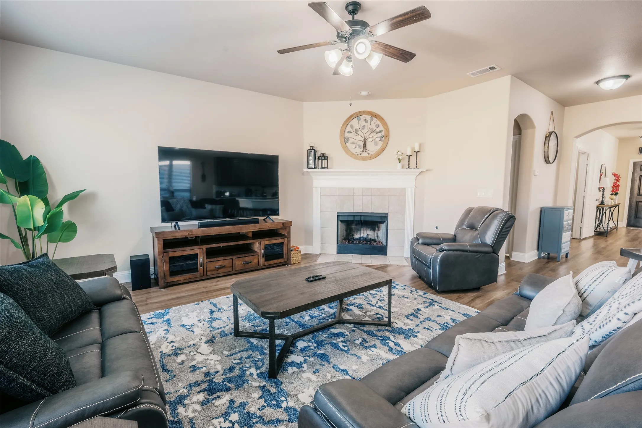 Spacious living area featuring a fireplace with tile surround, wood-finish flooring, a ceiling fan with integrated lighting, and arched doorways
