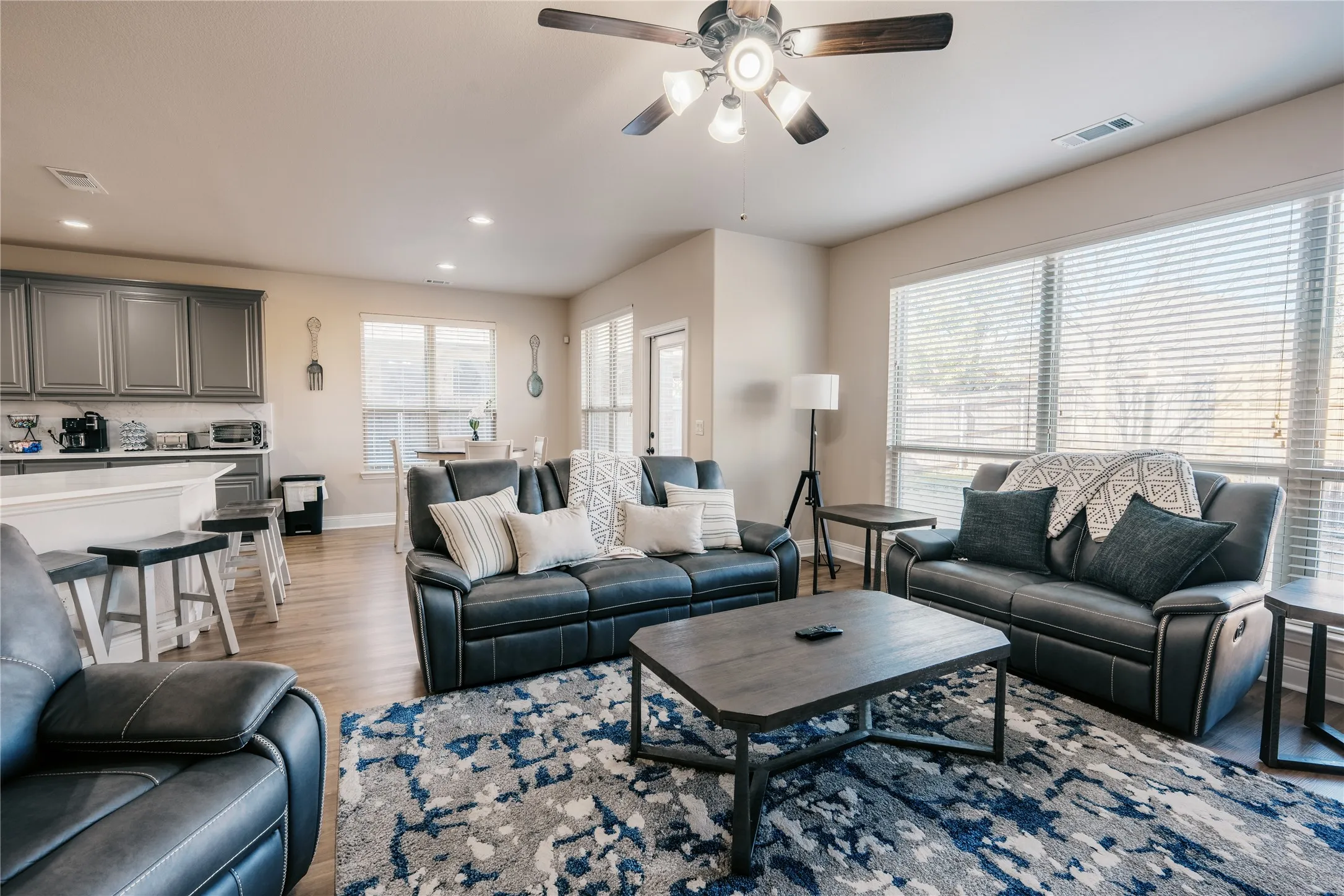 Open-concept living area featuring wood-finish flooring, a ceiling fan with integrated lighting, and multiple windows with blinds