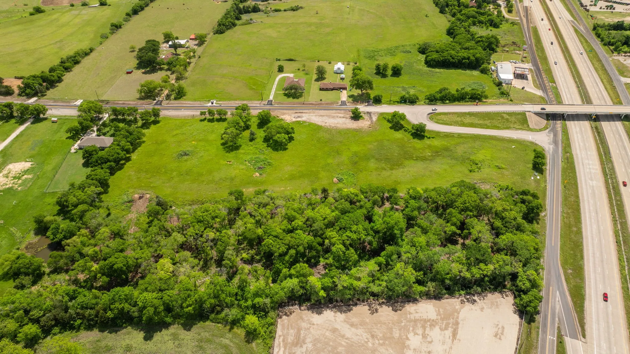 Expansive green acreage featuring a wooded section and multiple structures, bordered by a multi-lane highway with an overpass