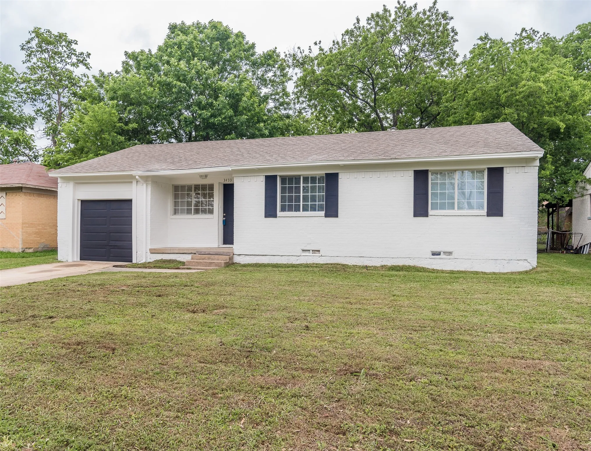 White brick exterior with dark shutters, an attached garage, a front entry door, and mature trees in the background