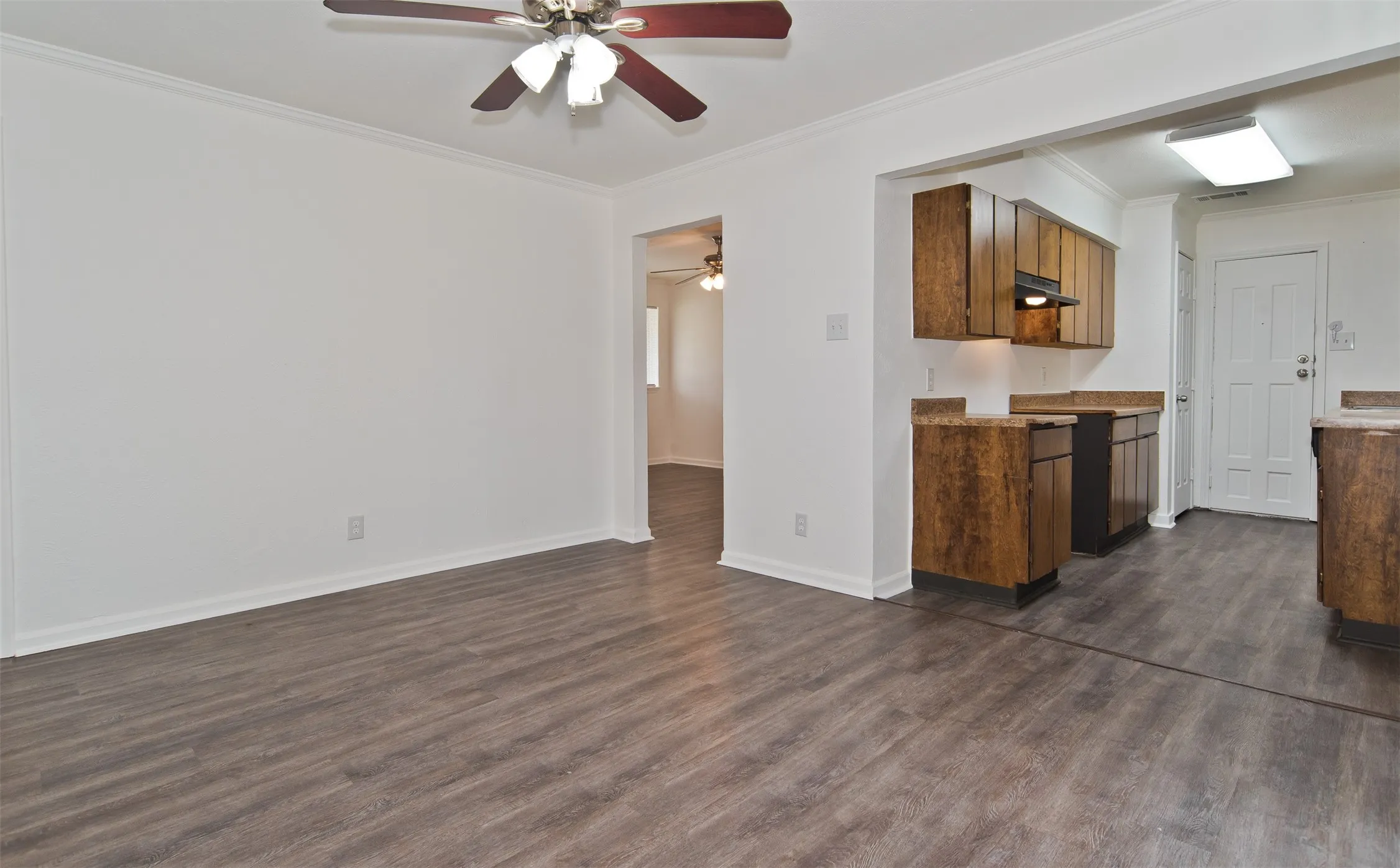Open concept living area with wood-finish flooring, a ceiling fan, crown molding, and an open doorway to the kitchen featuring wood cabinetry and countertops