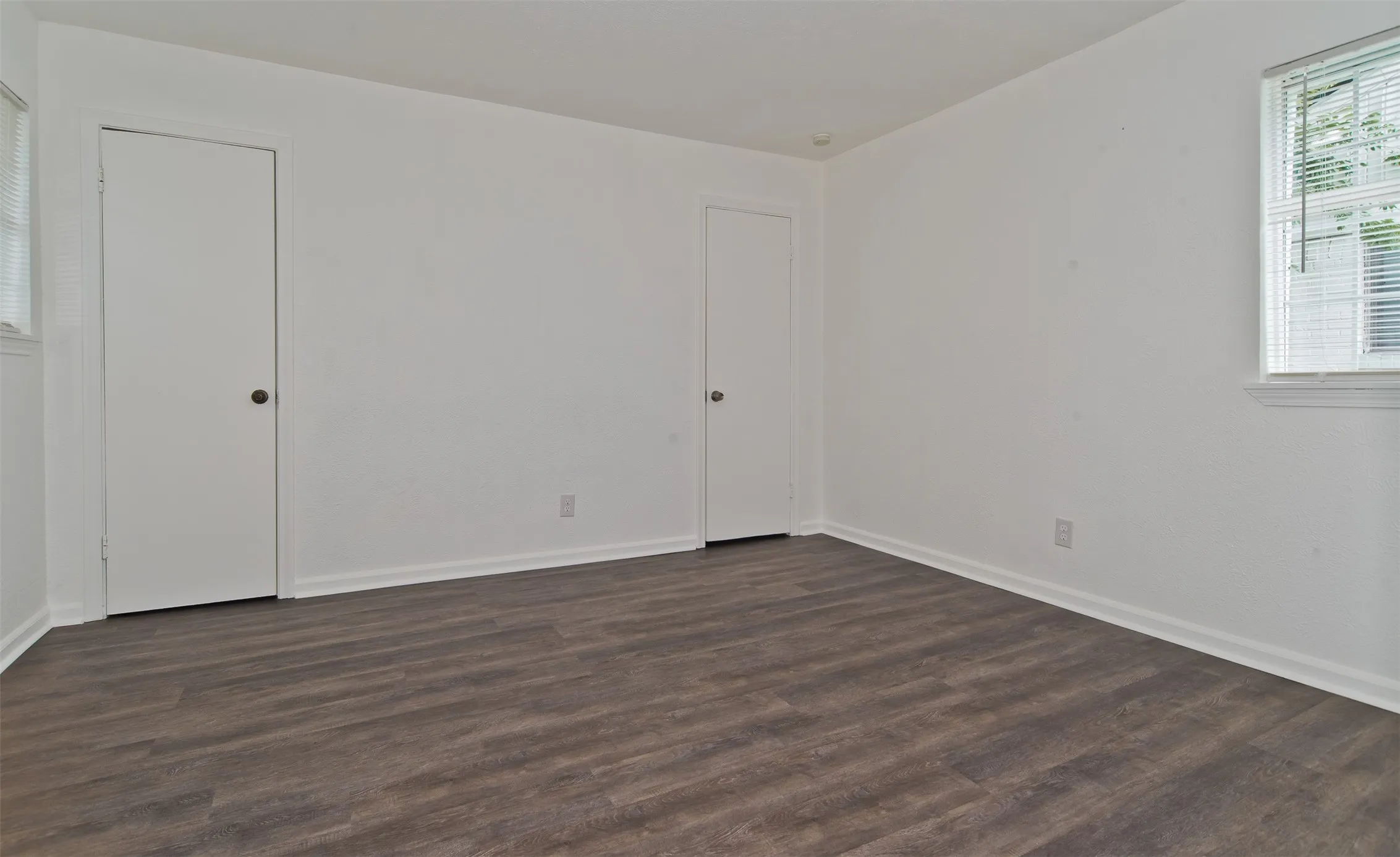 Interior room featuring wood-finish flooring, white walls, two white doors with dark hardware, and a window with blinds