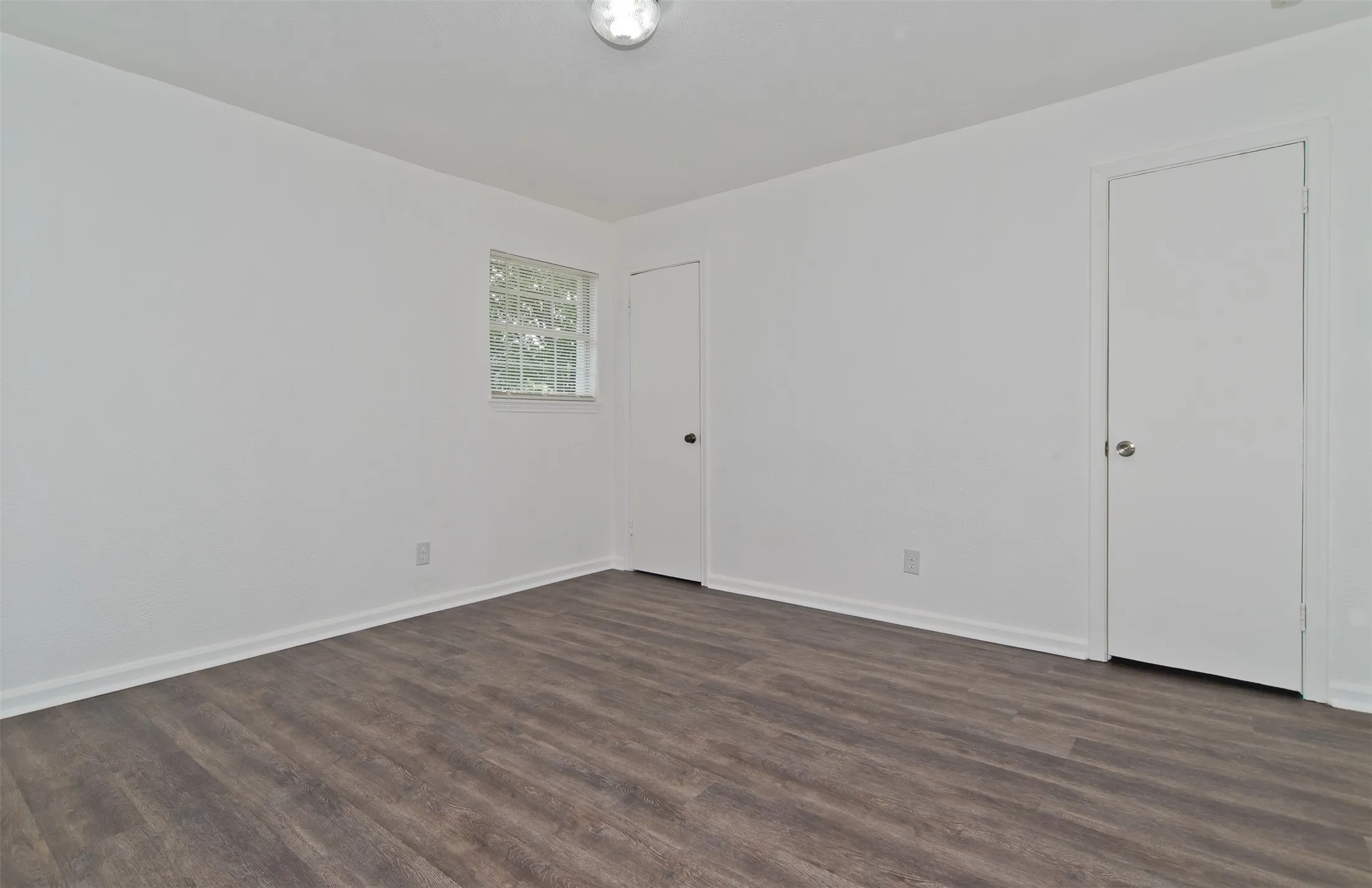 Spacious interior room featuring wood-finish flooring, white walls, and a ceiling-mounted light fixture