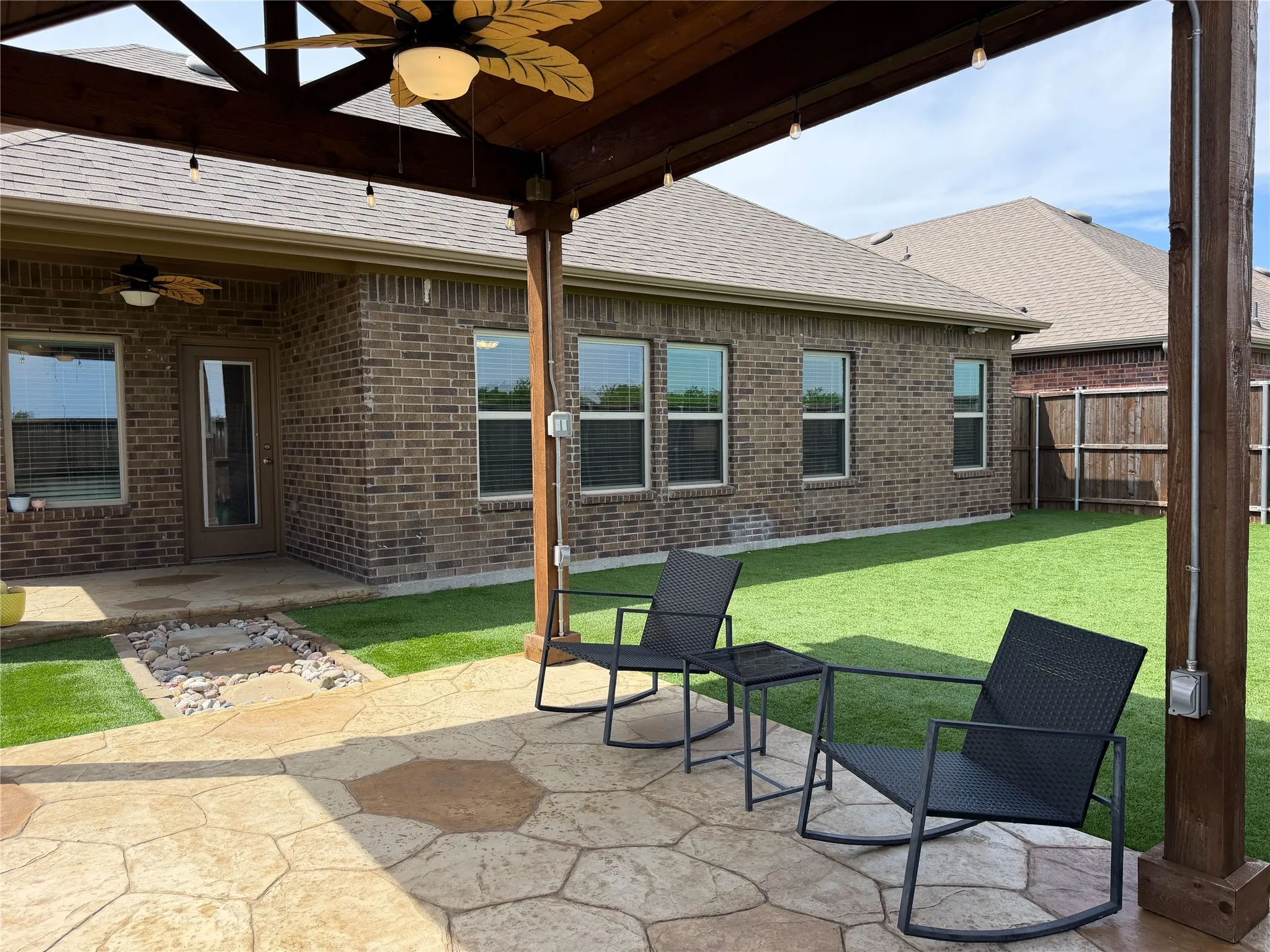 Step Outside to Enjoy Outdoor Living Space Flagstone and Rock Pathway Lead to 13 x 11 Artfully Crafted Pergola with a Solid Gabled Roof, Integrated Ceiling Fan, Bistro Lighting and Flagstone Inspired Stained Concrete. The Perfect Boutique Space for Year-Round Chilling. Easy to Maintain Artificial Grass in Back Yard.