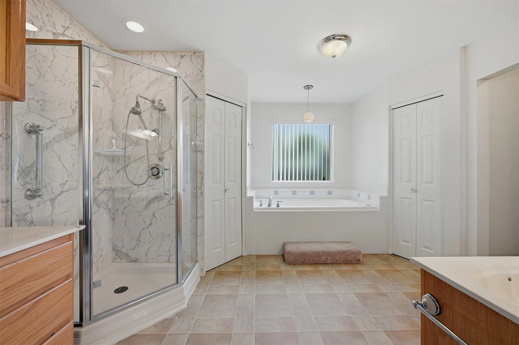 Spacious bathroom featuring a glass-enclosed shower with patterned wall tiling, a built-in soaking tub beneath a window, light wood-finish cabinetry, and neutral-toned tile flooring