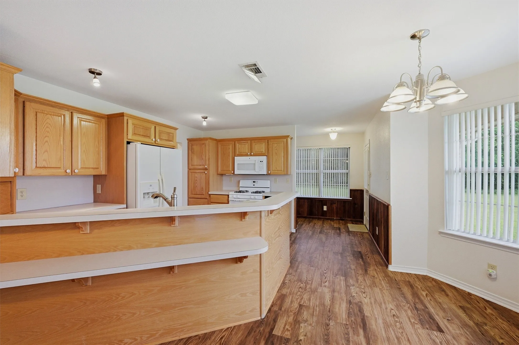 Kitchen and dining area featuring wood-finish flooring, light wood cabinetry, white appliances, and a breakfast bar