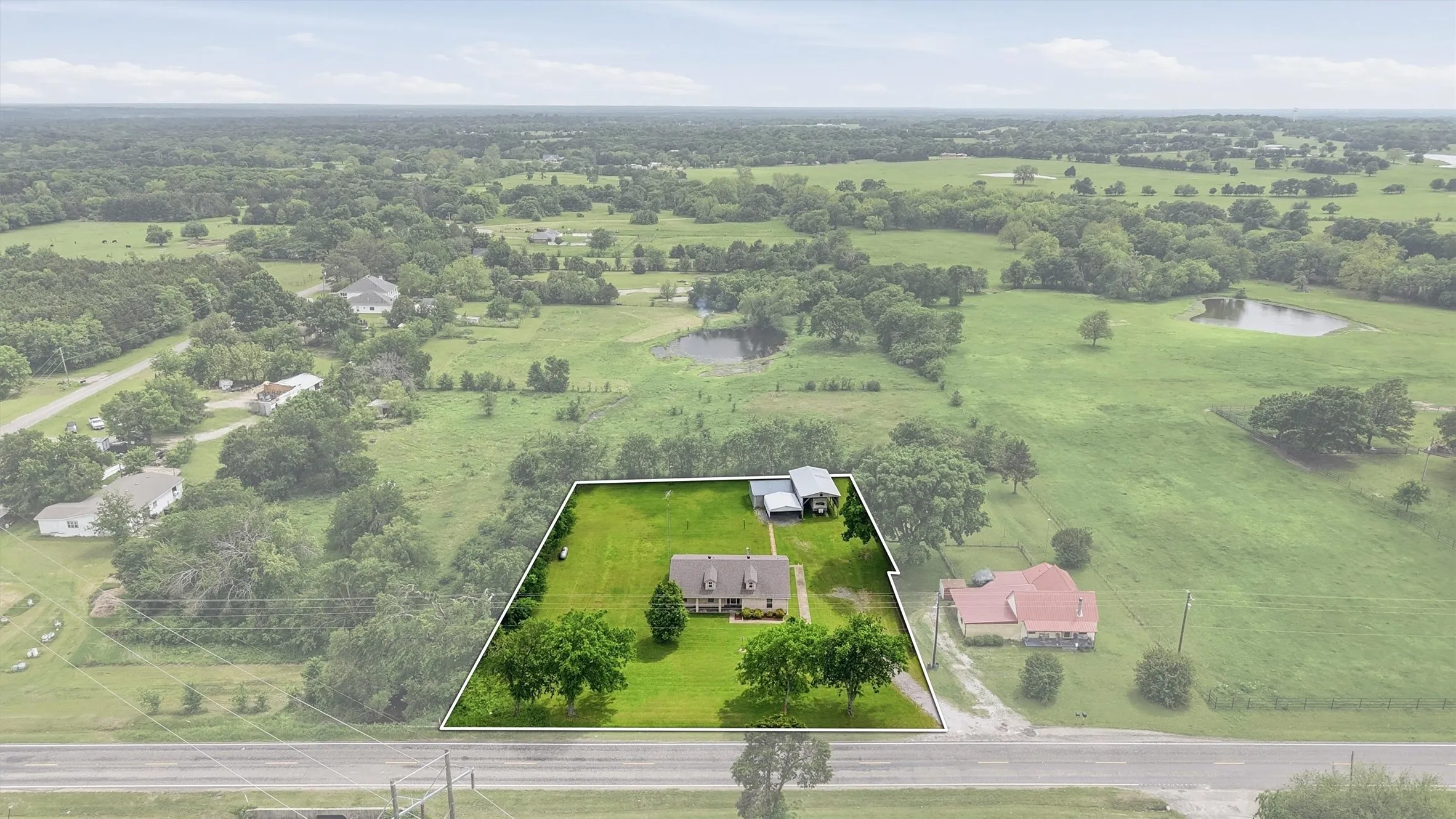 Expansive property featuring a single-story residence with a gabled roof, lush green lawn, mature trees, and an additional detached structure