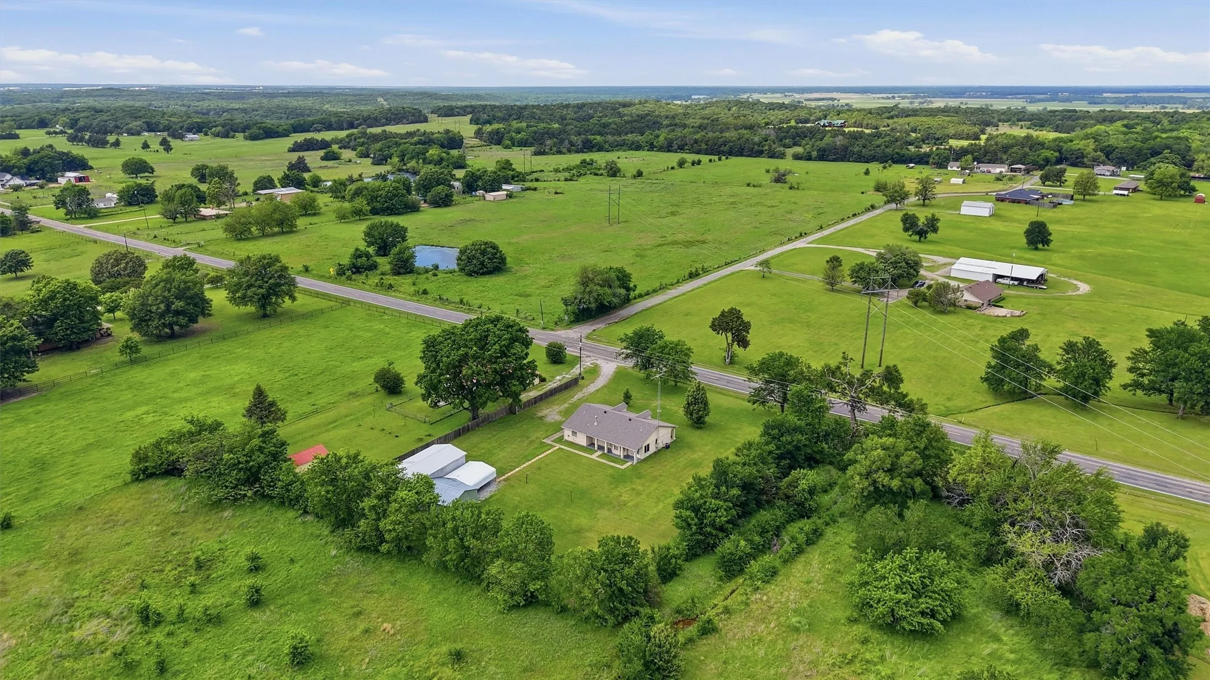 Expansive rural landscape featuring a single-story residence with an attached garage, multiple outbuildings, and a surrounding green field