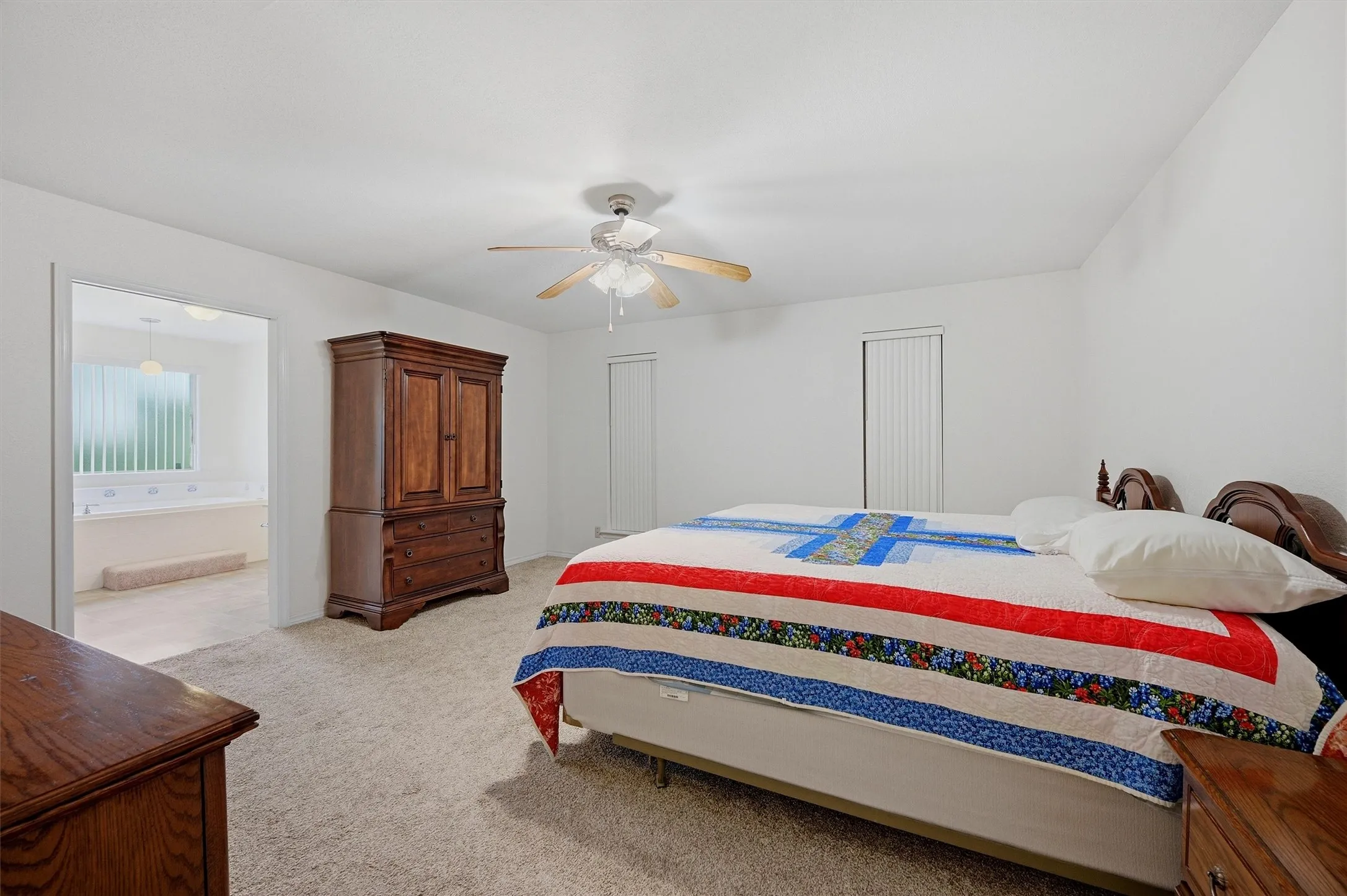Carpeted room featuring a ceiling fan, white walls, and a doorway opening to a bathroom with a built-in tub and privacy window