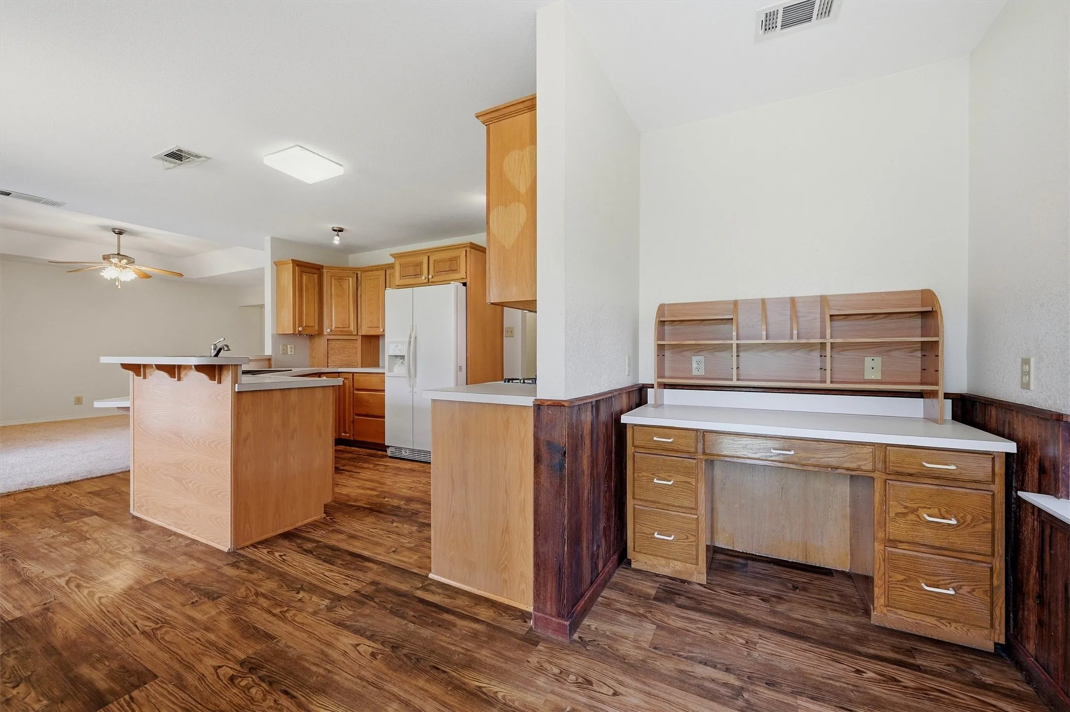 Kitchen and built-in desk area featuring wood-finish flooring, light wood cabinetry, white countertops, a central island with a sink, and a ceiling fan in the background