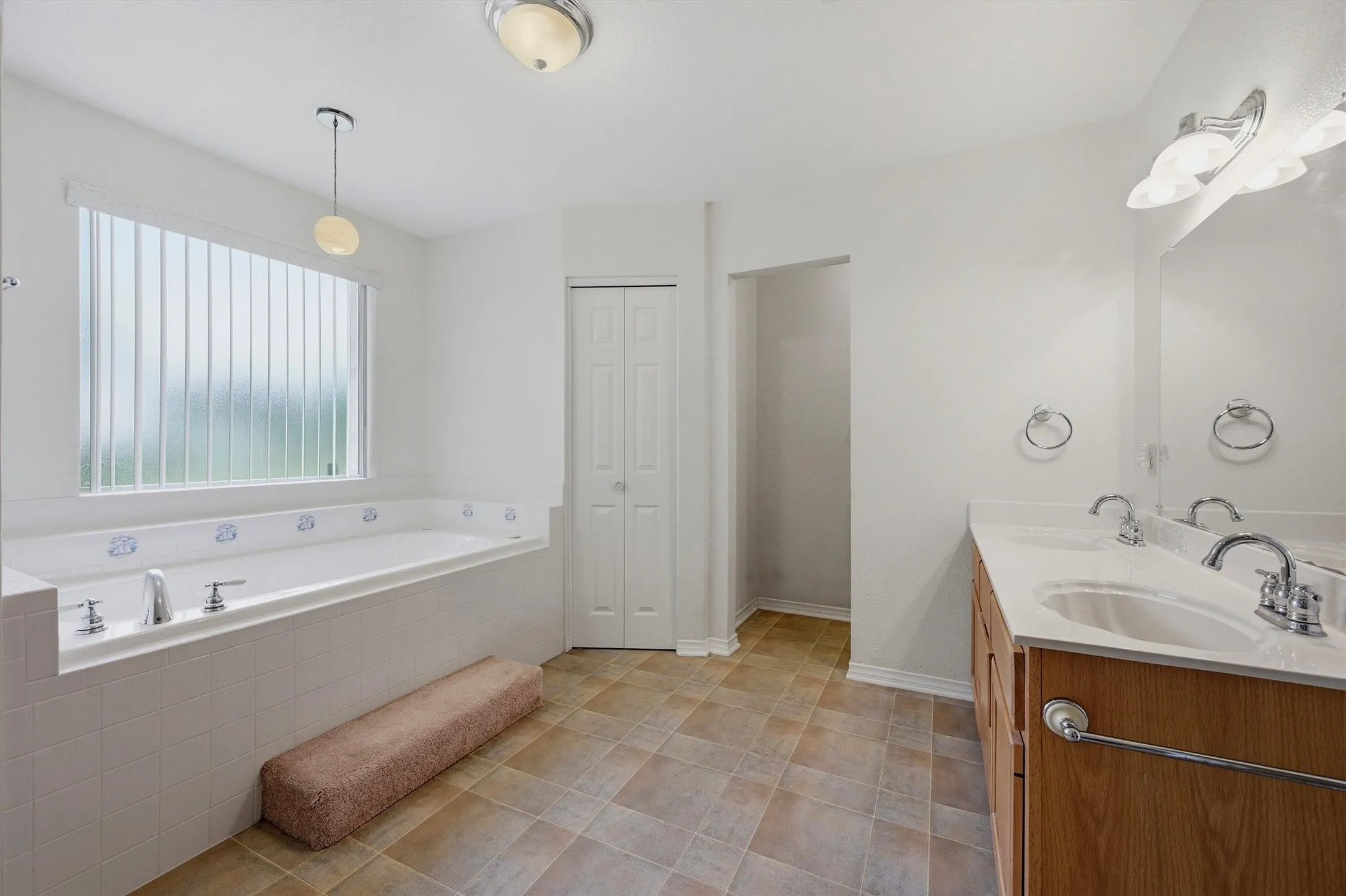 Spacious bathroom featuring a built-in soaking tub with chrome fixtures, a large window with vertical blinds, a double vanity with an integrated mirror, and patterned tile flooring