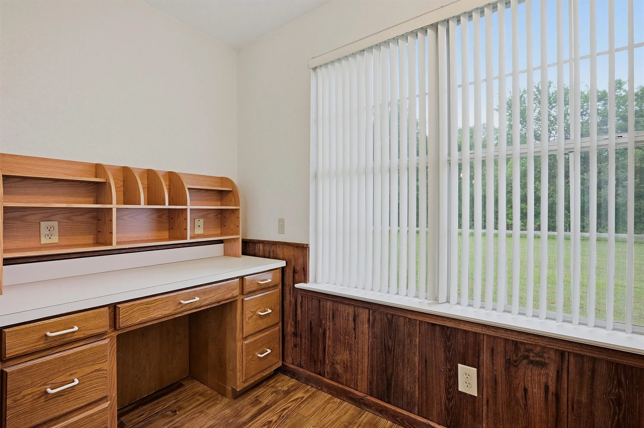 Room featuring a built-in wood desk with overhead shelving, a large window with vertical blinds, wood-finish flooring, and wood paneling on the lower wall