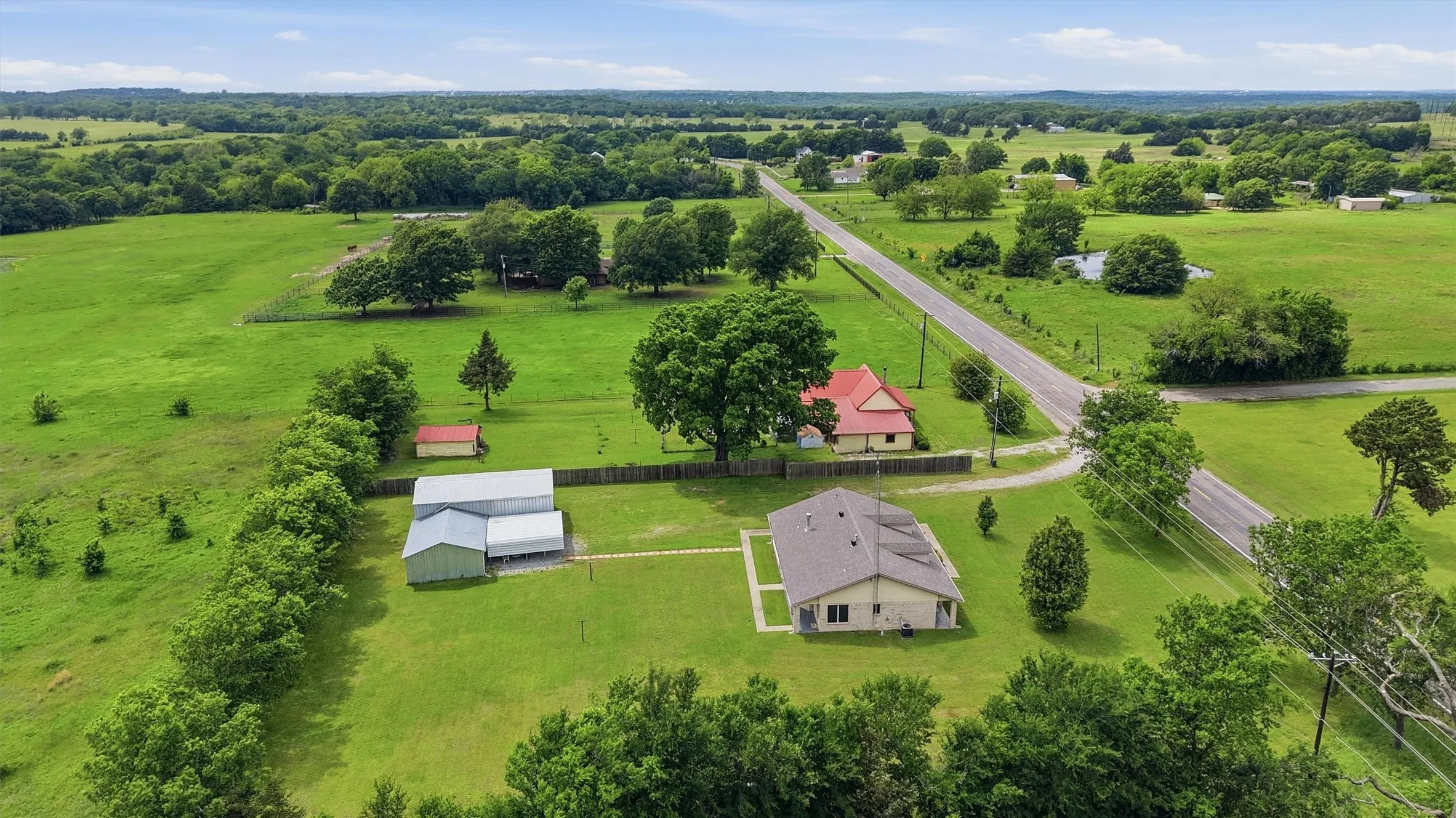 Expansive rural property featuring a single-story residence with a gray shingle roof, green grass landscaping, two large outbuildings, a small red storage shed, and mature trees