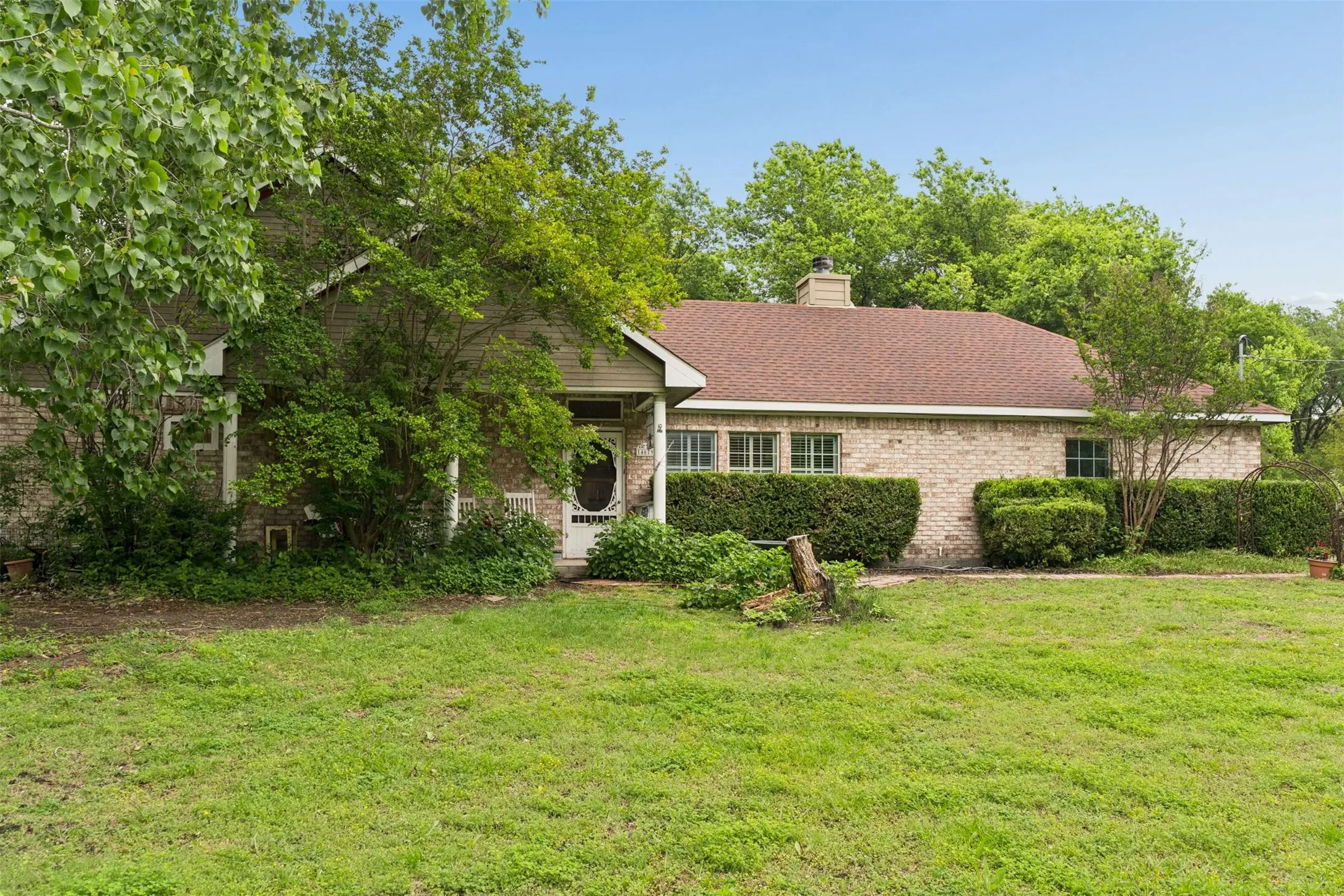 Traditional style brick residence with a brown shingle roof, featuring multiple windows, a front porch, and mature landscaping