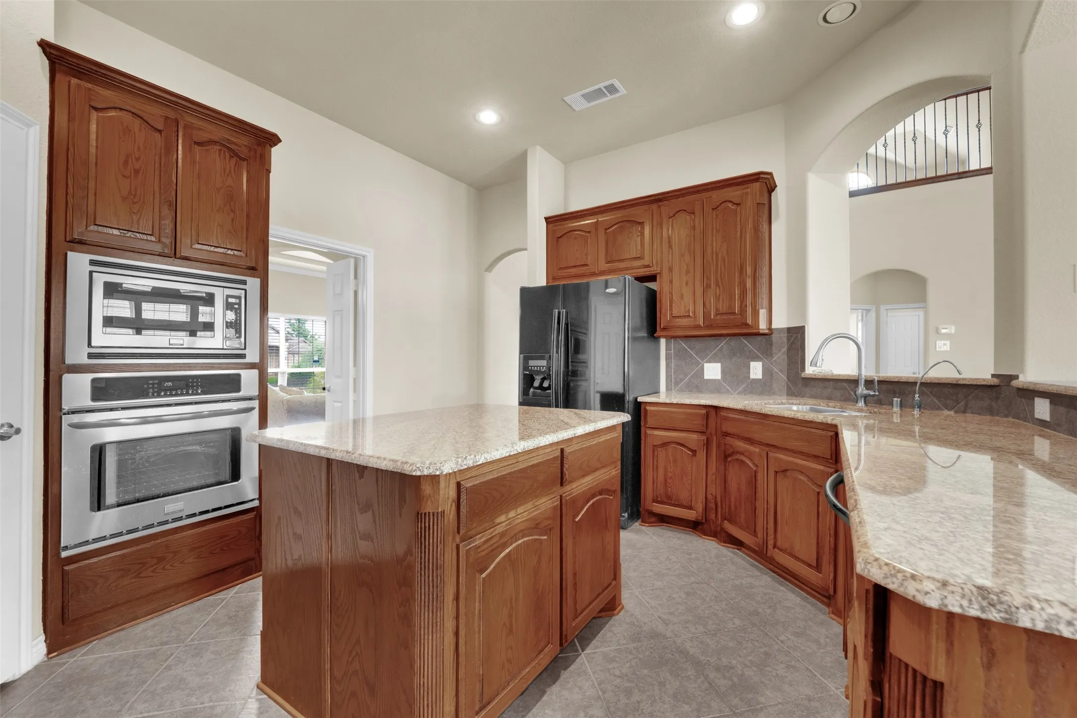 Kitchen featuring a central island with a light-toned granite countertop, a full suite of stainless steel appliances including a built-in oven and microwave, extensive wood cabinetry, and a tile backsplash