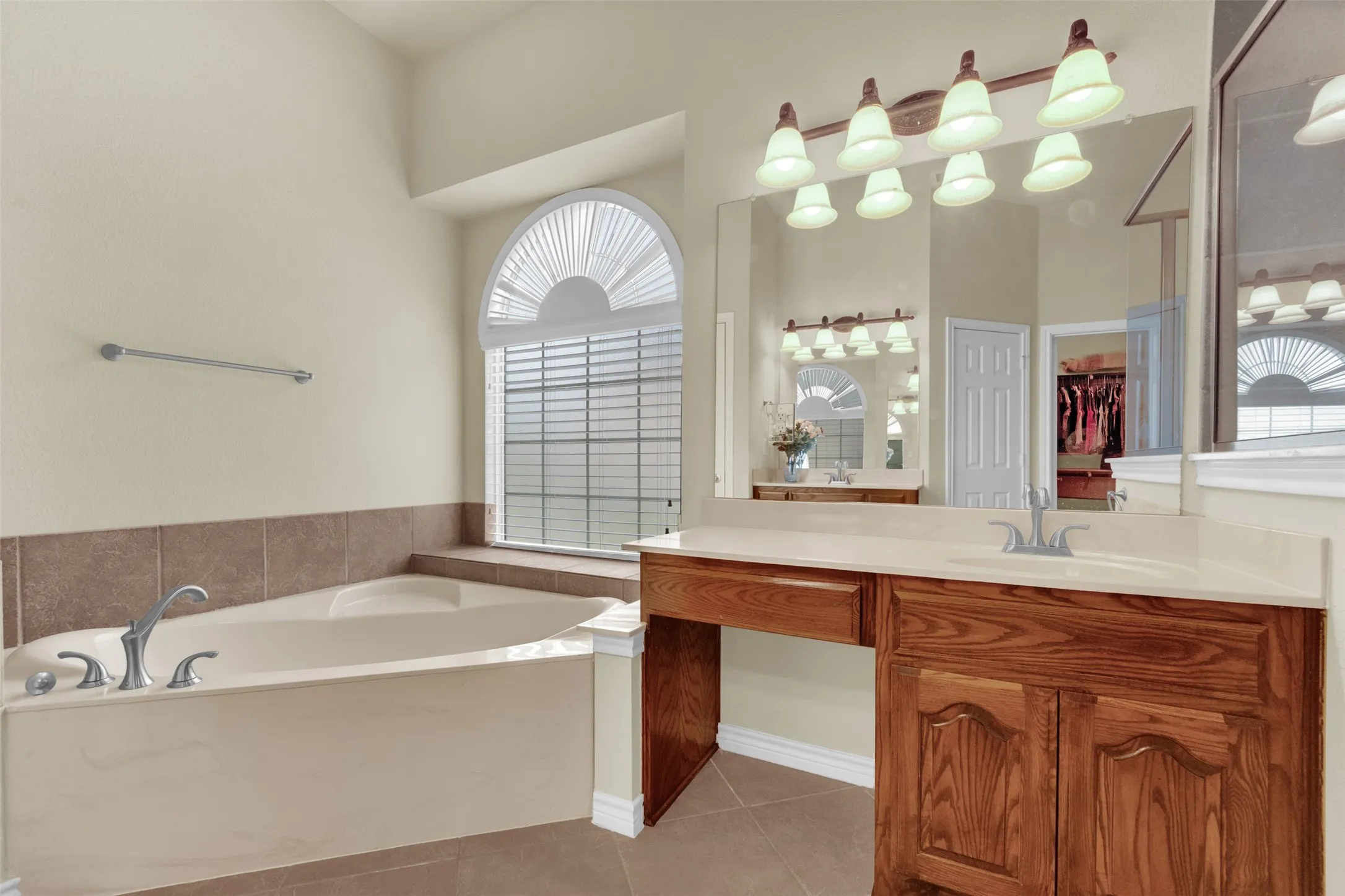 Bathroom featuring a corner soaking tub with deck-mounted fixtures, an arched window with blinds, a wood-finish vanity with countertop, and an expansive wall-mounted mirror with overhead lighting