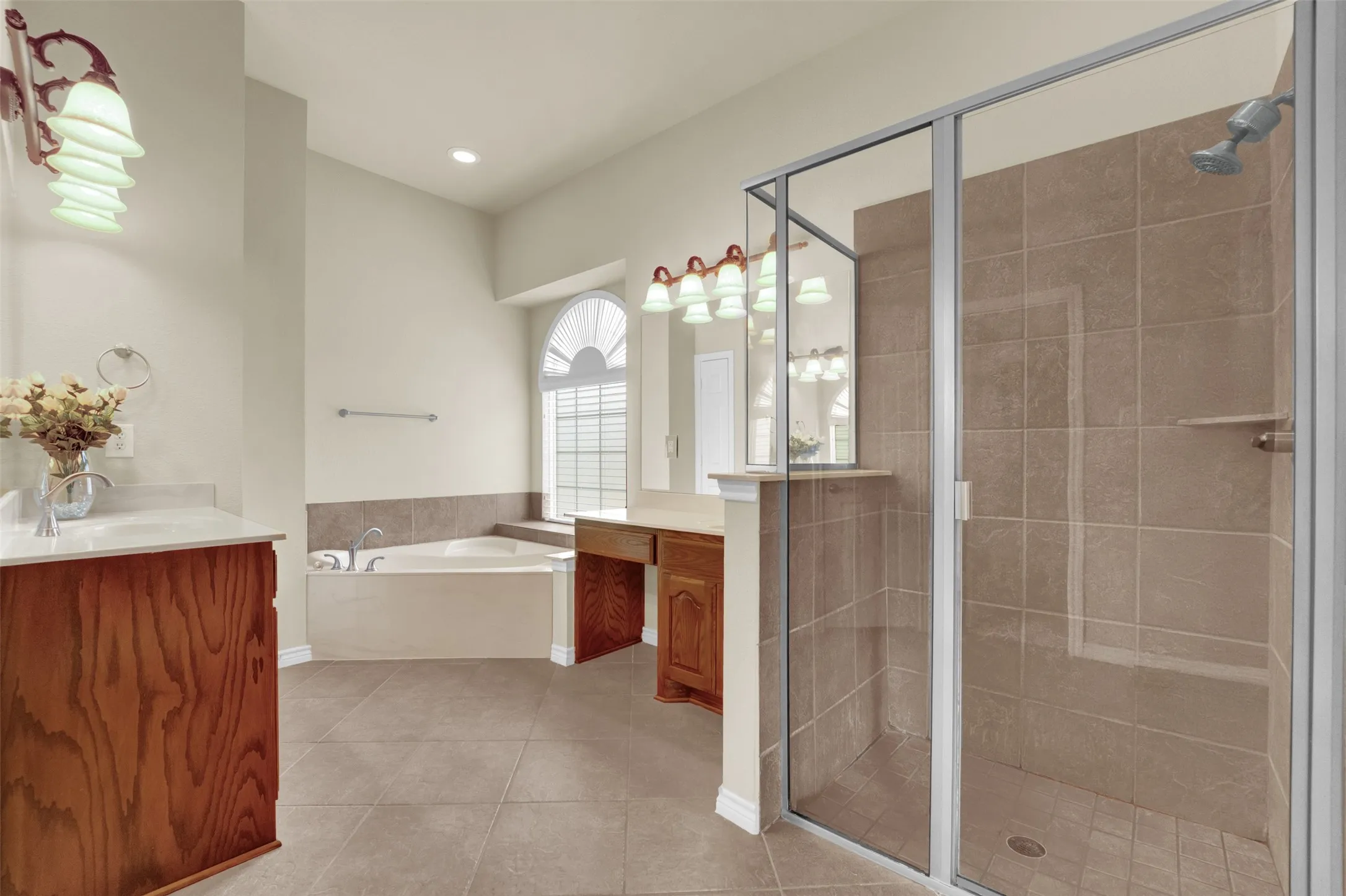 Spacious bathroom featuring a glass-enclosed shower, corner soaking tub, dual vanity areas with wood-finish cabinetry, and an arched window with louvered blinds