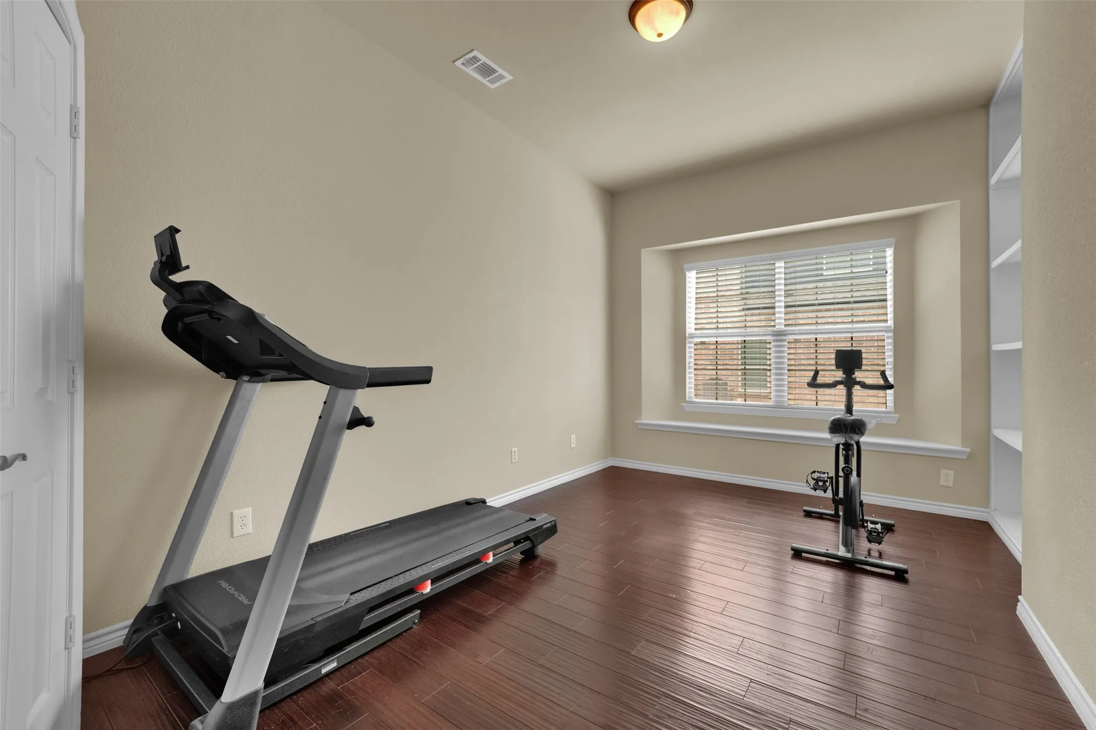 Spacious room featuring rich wood-finish flooring, a large window with blinds, and a built-in shelving unit