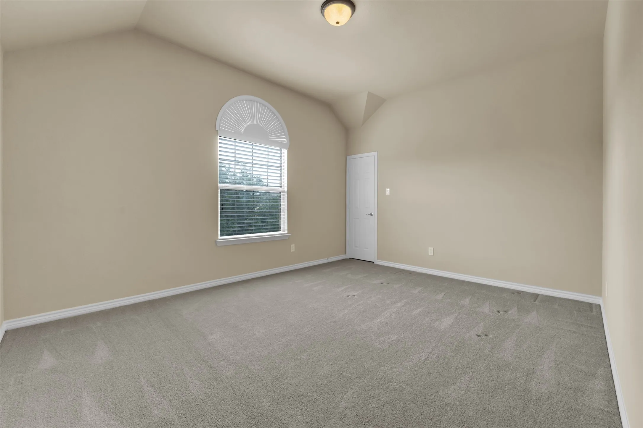 Spacious room featuring vaulted ceilings, light-toned carpeting, a large arched window with blinds, and neutral wall paint