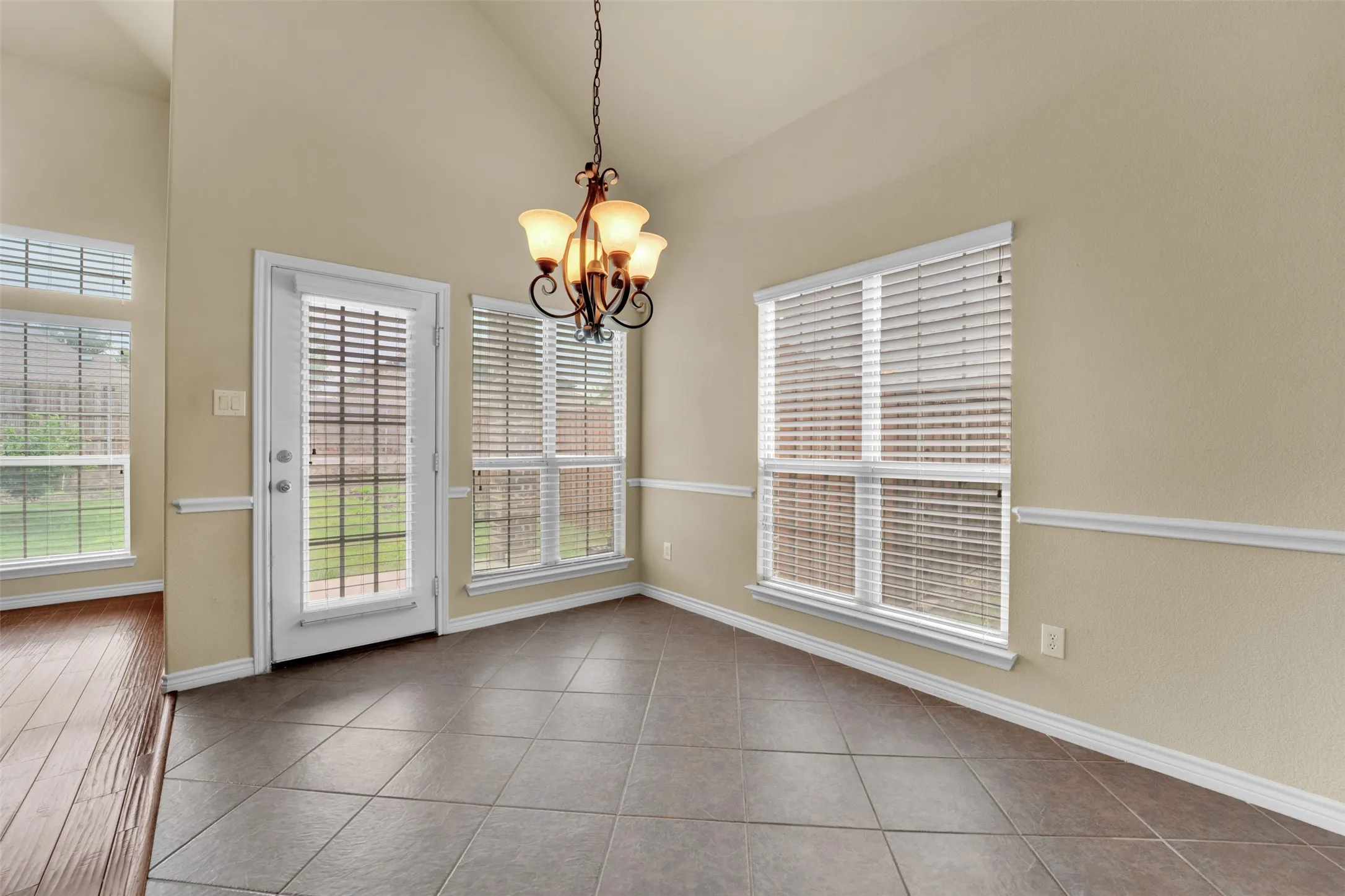 Dining area with vaulted ceiling, featuring tile flooring, multiple windows with blinds, a full-lite door, and an ornate chandelier