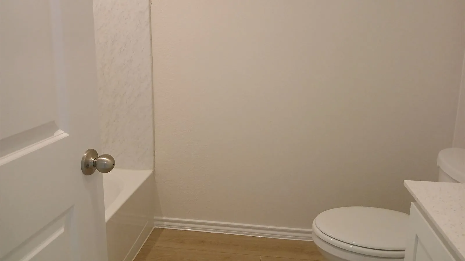 White paneled door with brushed nickel hardware, built-in bathtub, light-toned wall finish, white toilet, and wood-finish flooring