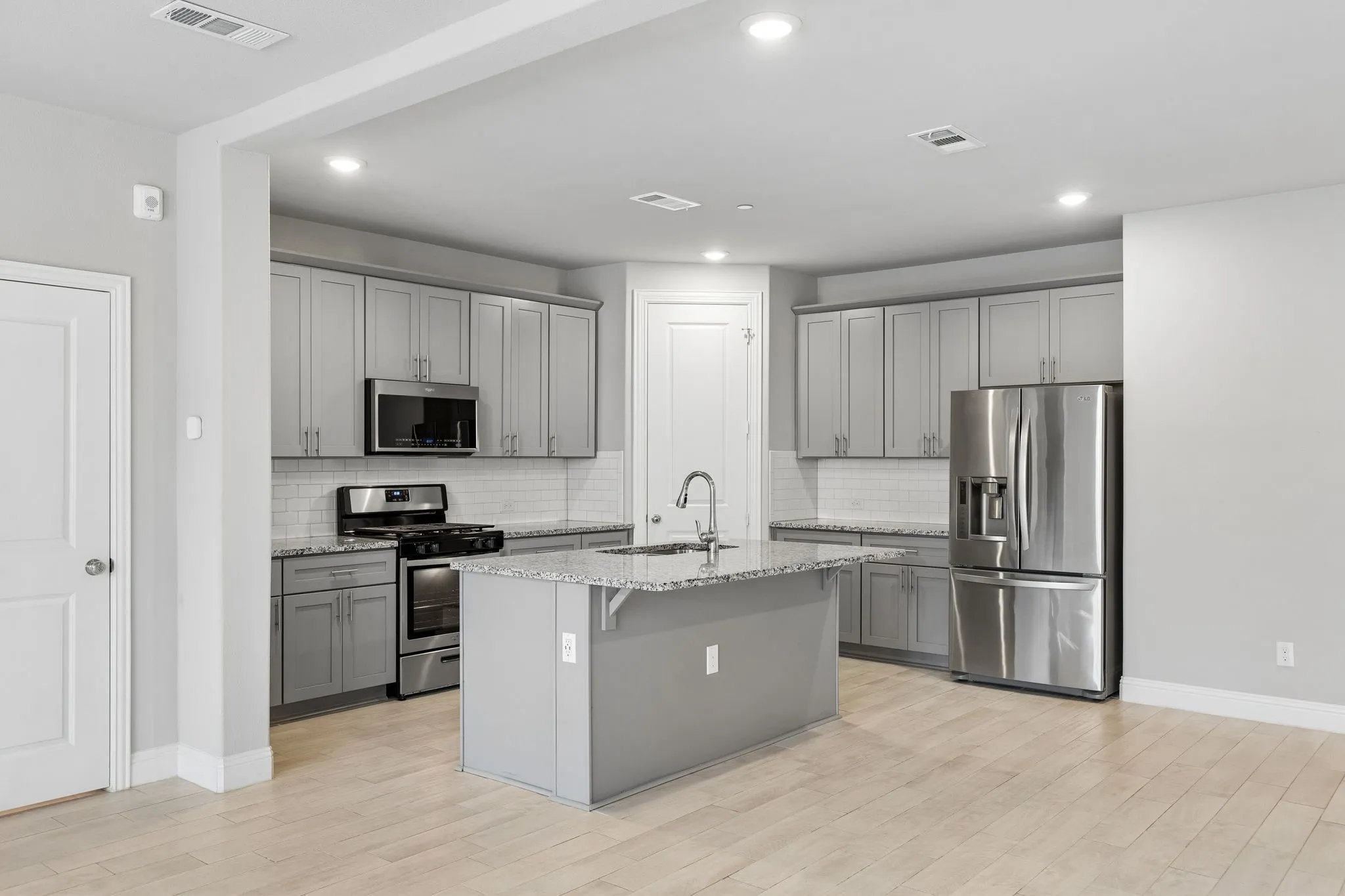 Modern kitchen featuring light gray cabinetry, granite counter tops, stainless steel appliances, a center island with a sink, light-toned wood-finish flooring, and recessed lighting