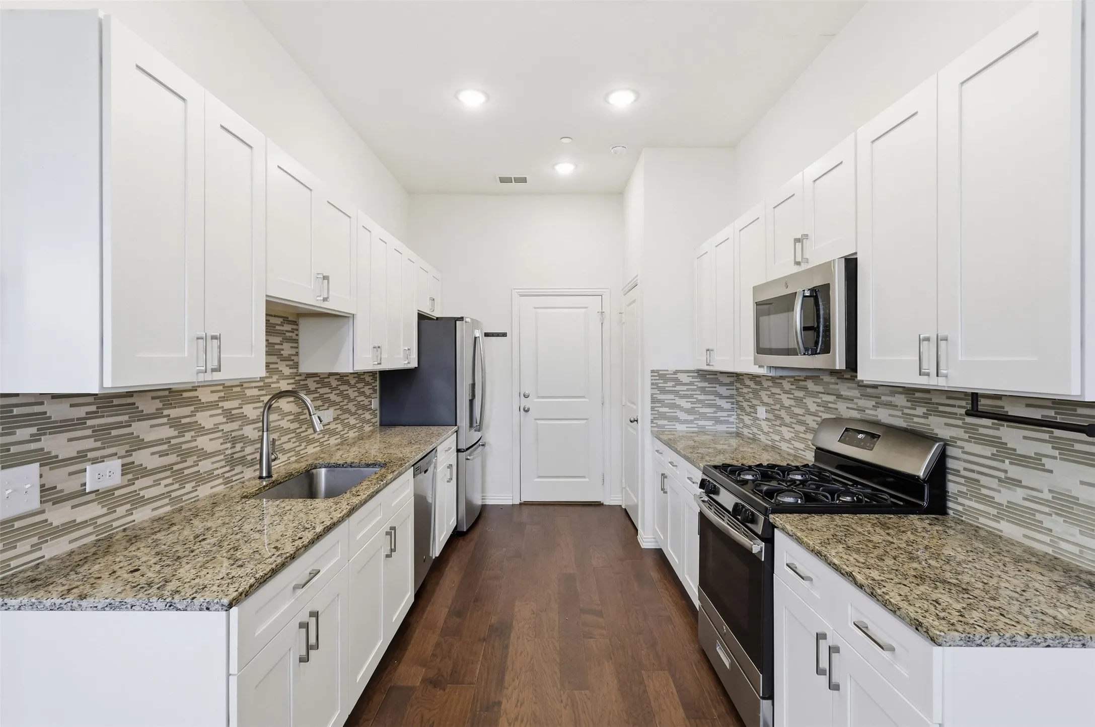 Galley kitchen with white shaker-style cabinetry, stainless steel appliances, granite countertops, and a mosaic tile backsplash. Fridge included