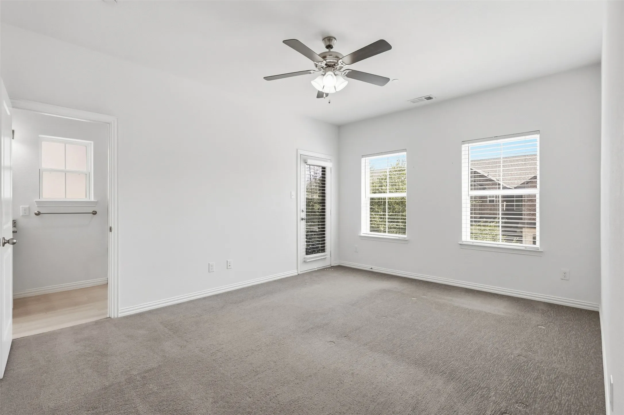 Spacious room featuring carpet flooring, a ceiling fan with integrated lighting, two windows with blinds, and a glass-paneled exterior door to private balcony.