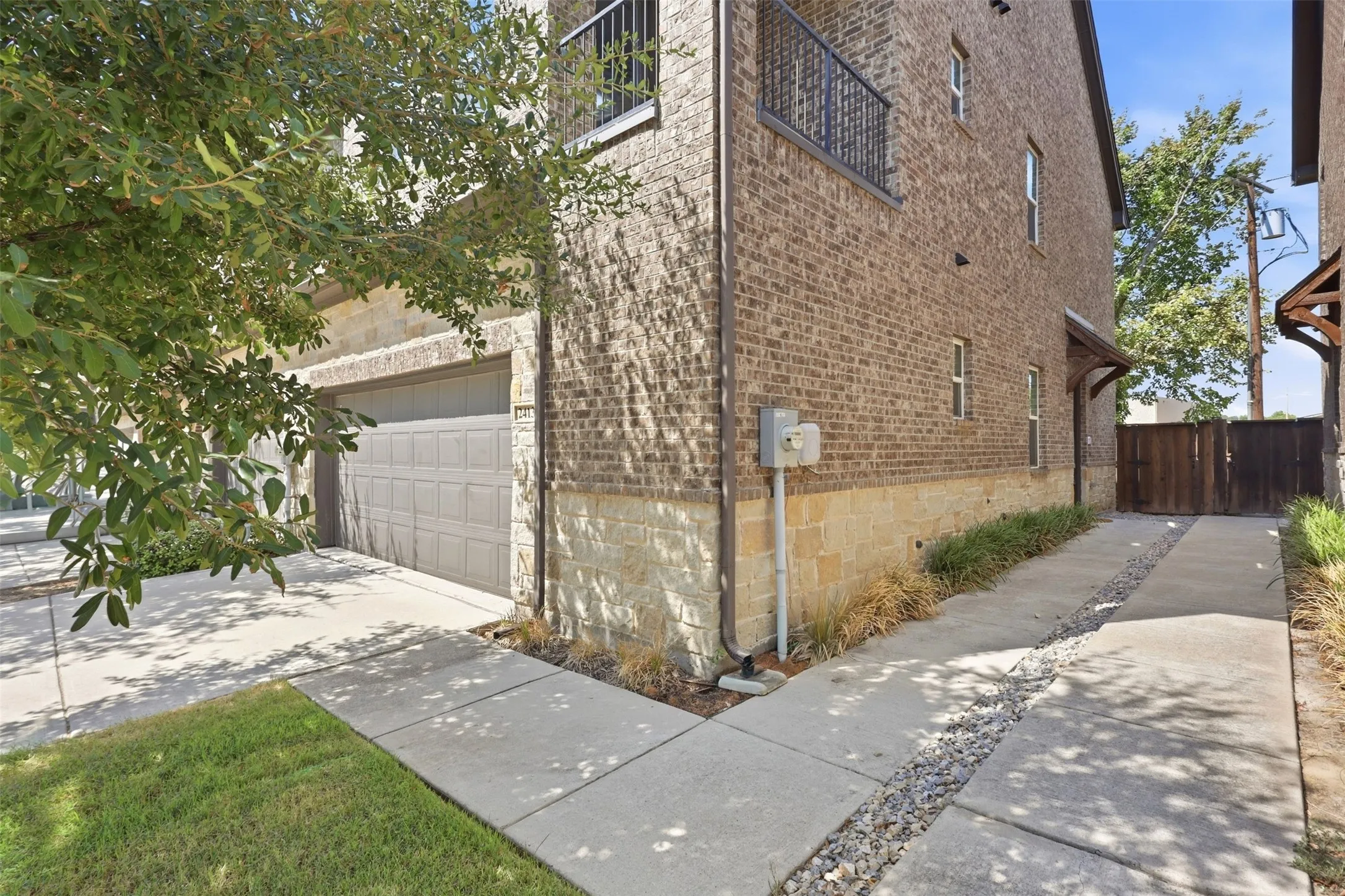 Brick and stone facade, wrought iron balcony, paneled garage door, concrete driveway, and natural wood fencing with new landscaping along entryway