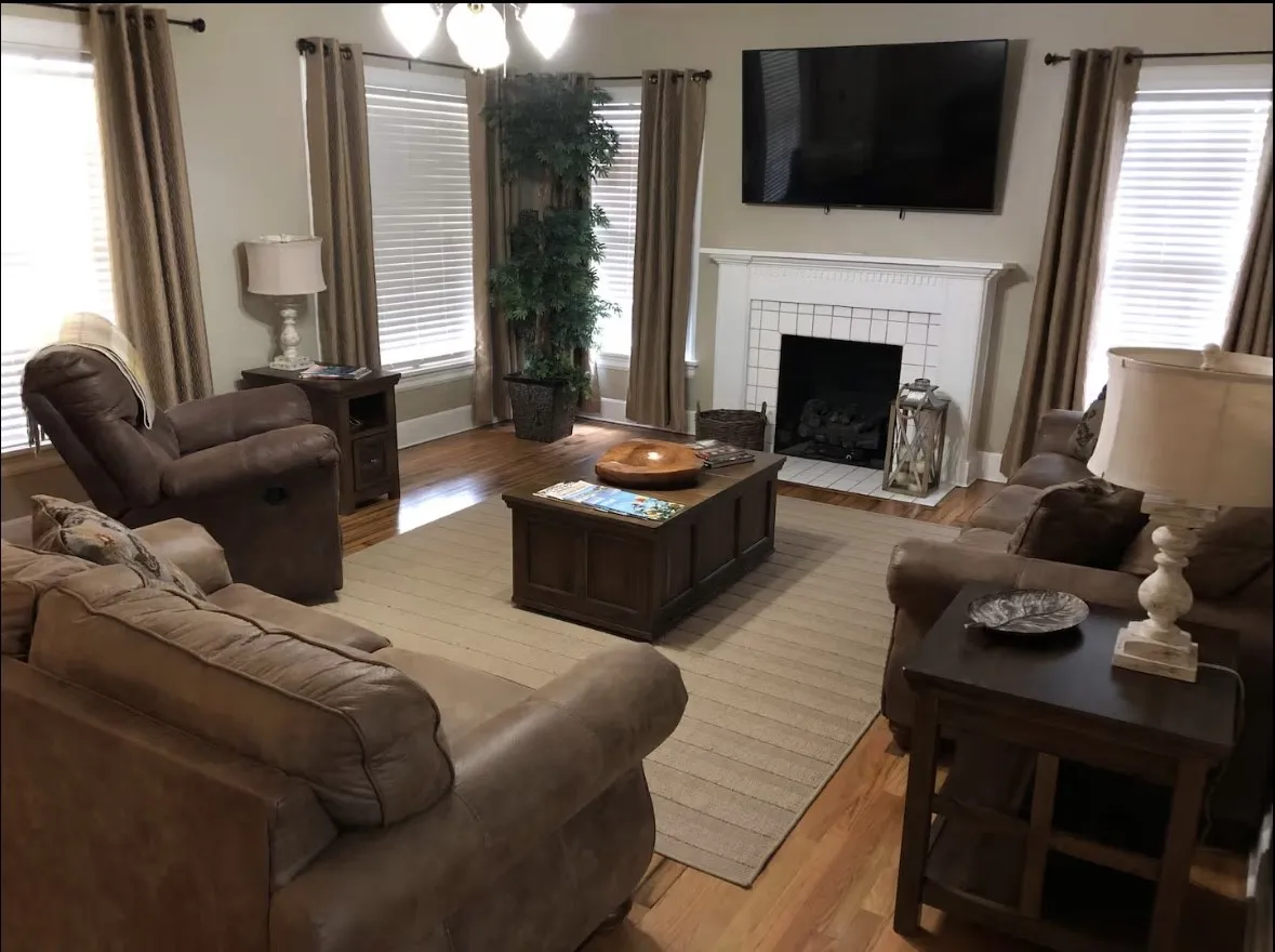 Living space featuring wood-finish flooring, multiple large windows with blinds, a prominent fireplace with a white mantel, and a ceiling fan with integrated lighting