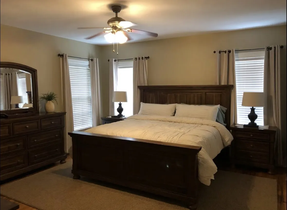 Spacious bedroom featuring warm-toned walls, a ceiling fan with integrated lighting, two windows with blinds and drapes, and a combination of carpet and wood-finish flooring