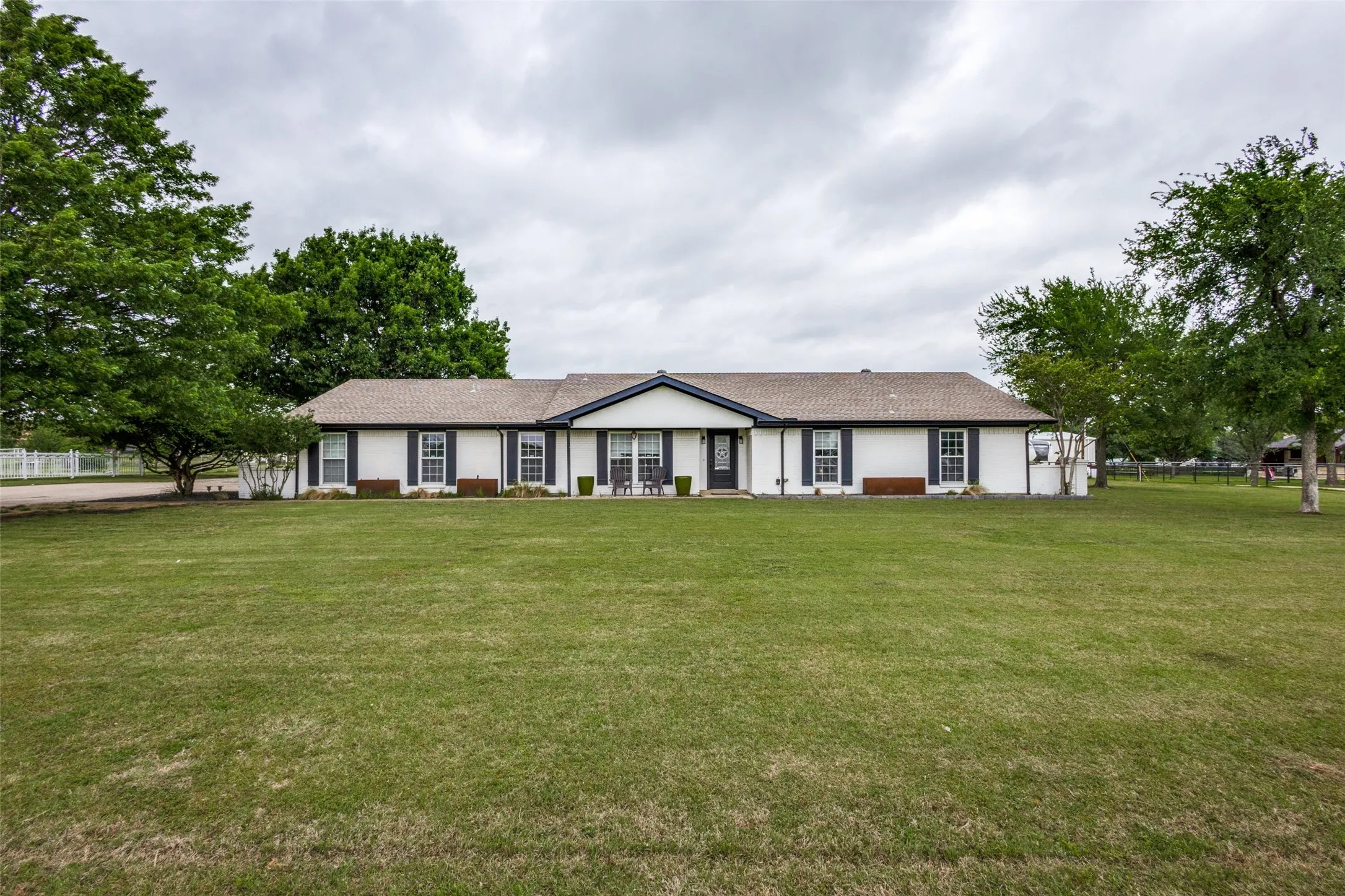 Spacious front lawn with a single-story white facade residence featuring dark shutters, a covered entry, and mature trees