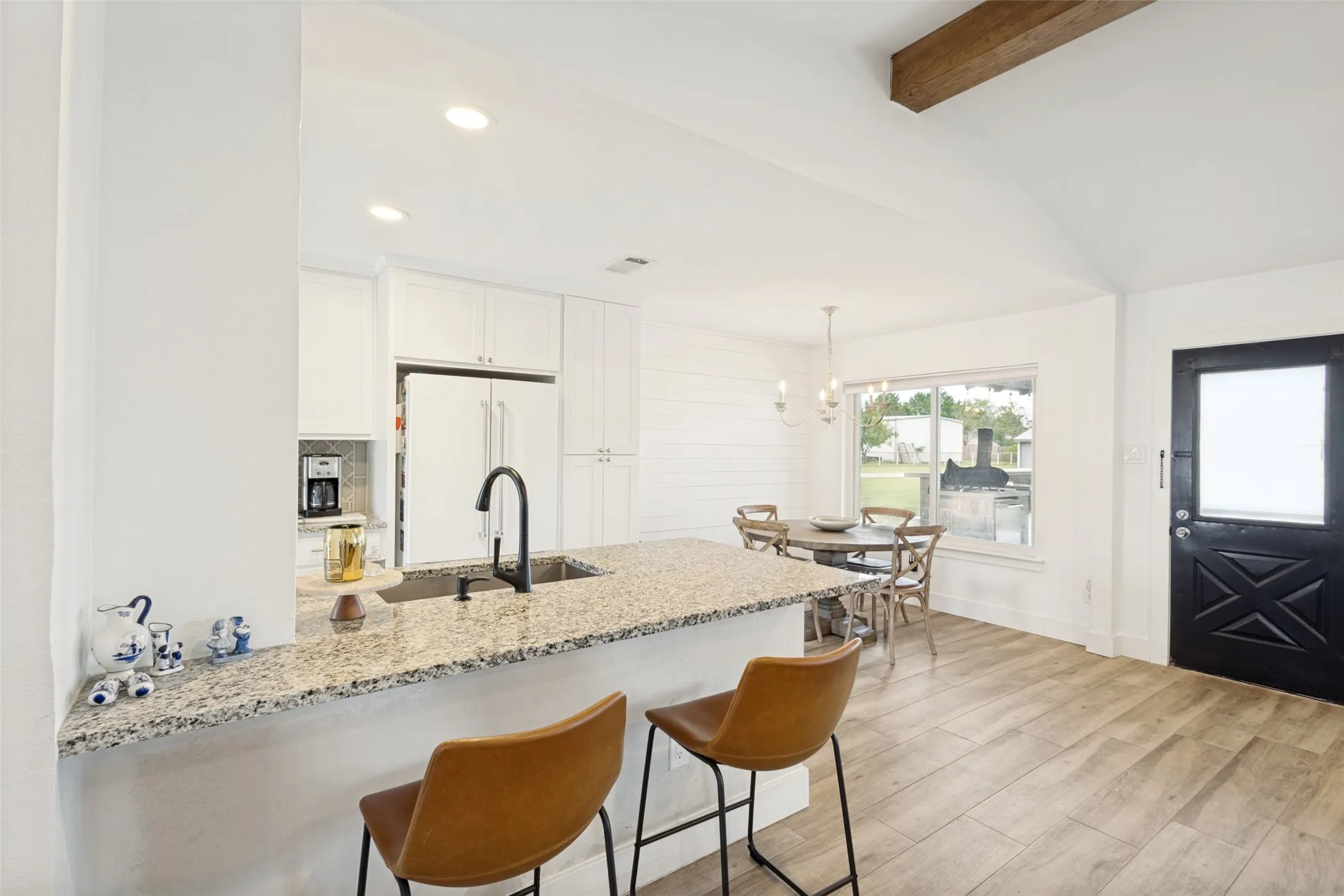 Open-concept kitchen and dining area featuring a vaulted ceiling with a wood beam, granite countertops, white cabinetry, wood-finish flooring, and a black paneled door